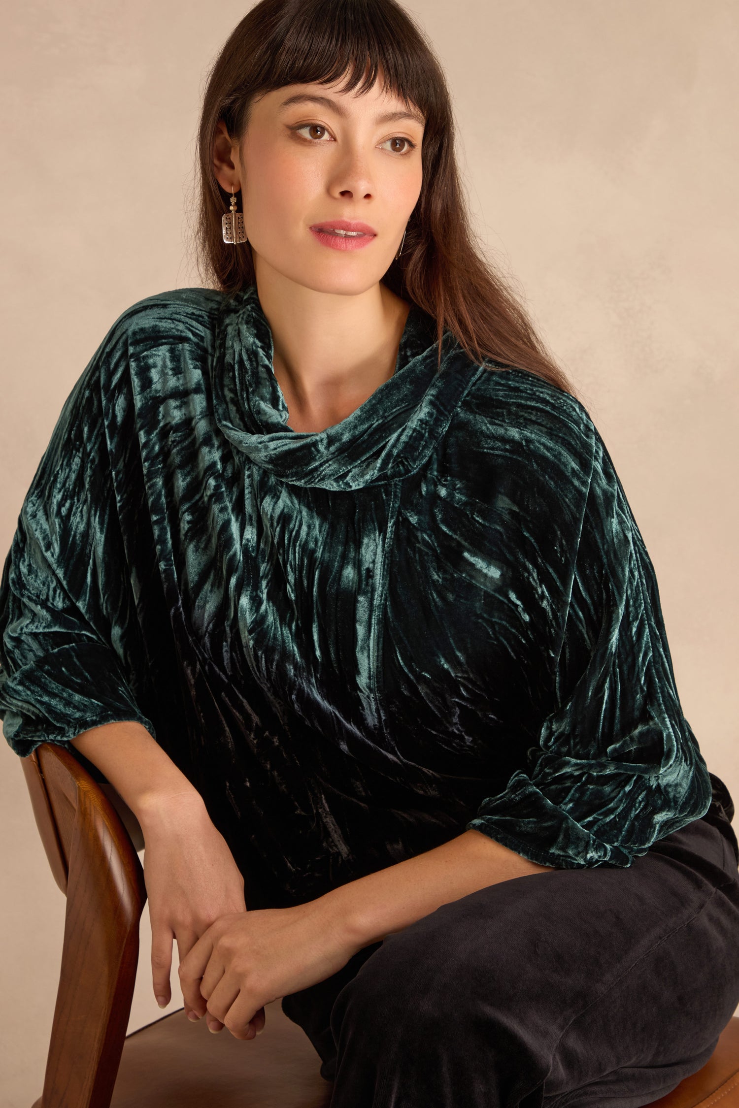Woman with long brown hair sits on a wooden chair, wearing the Turquoise Silk Velvet Ombre Cowl Neck Top and black pants against a neutral background.