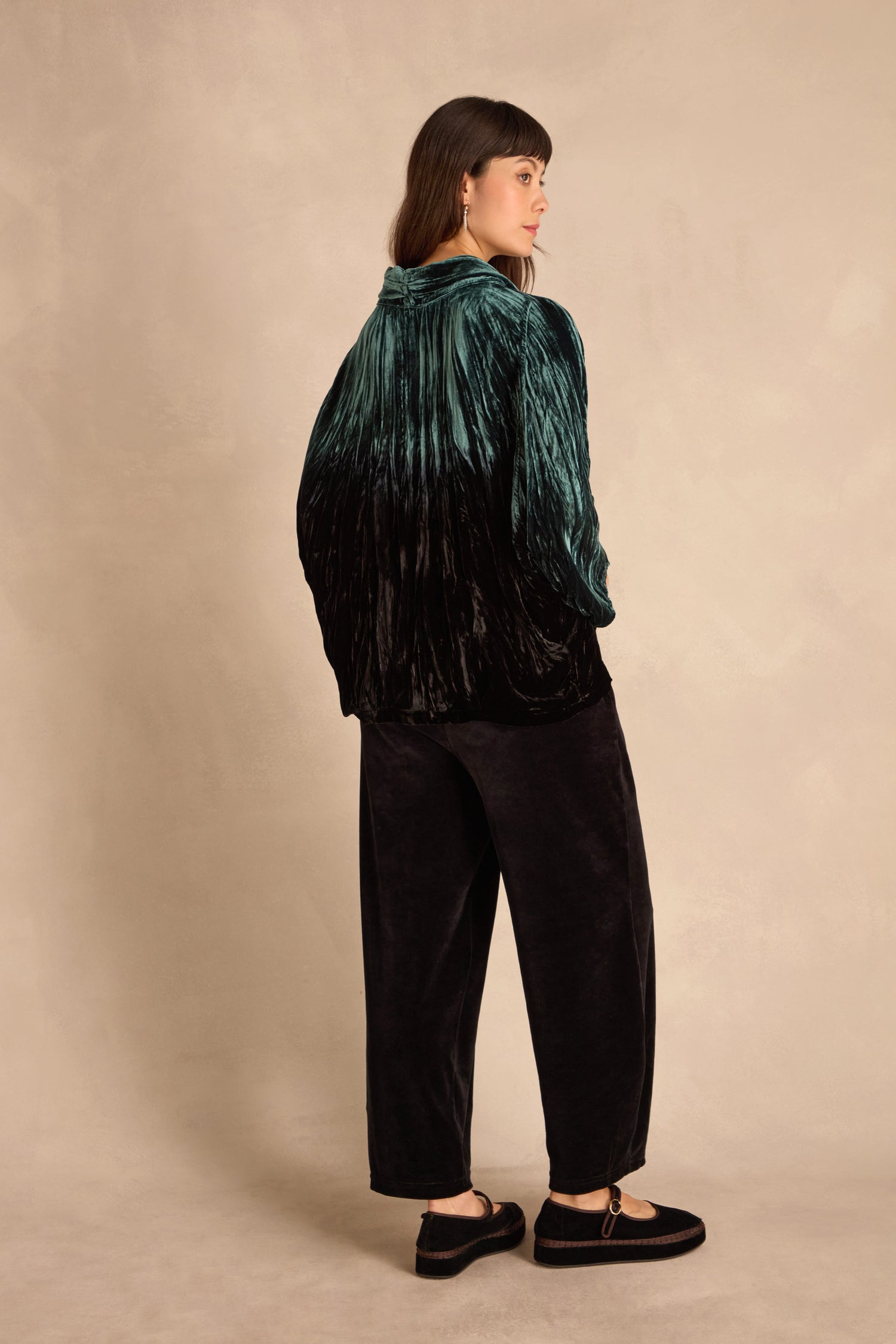 Woman standing and facing away, wearing a Turquoise Silk Velvet Ombre Cowl Neck Top, black wide-leg pants, and black shoes against a beige background.