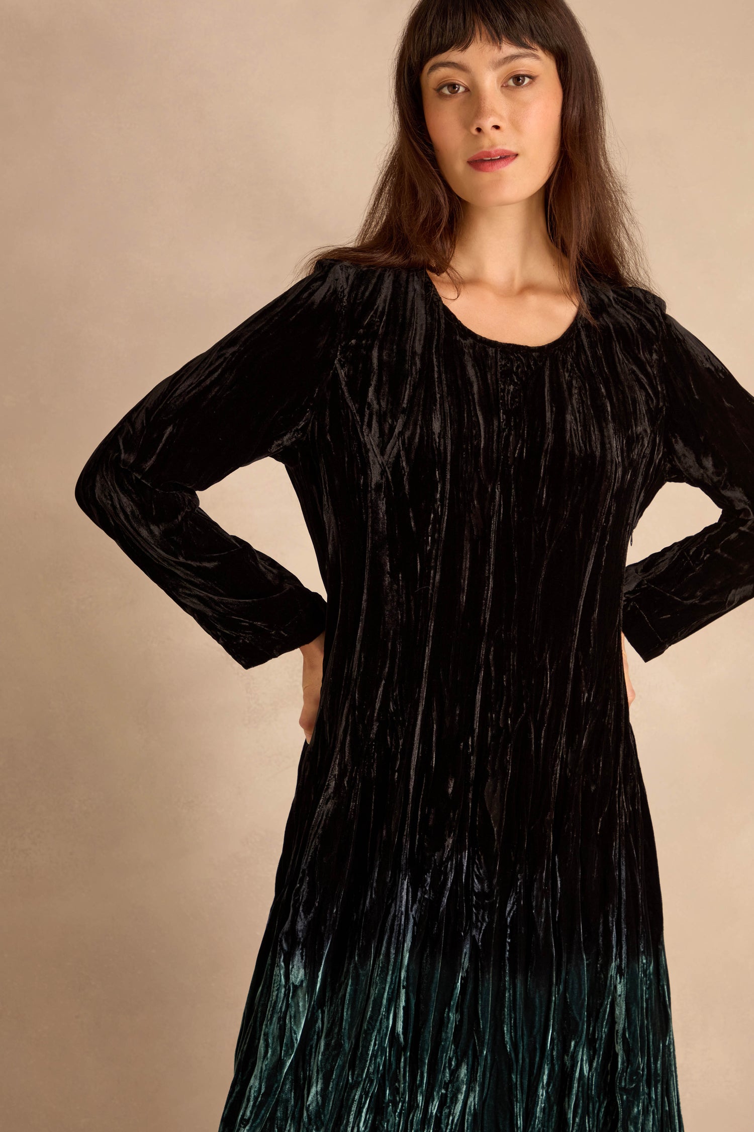 Woman poses hands on hips in the Silk Velvet Ombre Bubble Dress, featuring long sleeves and a hem that fades into dark green, against a neutral background.
