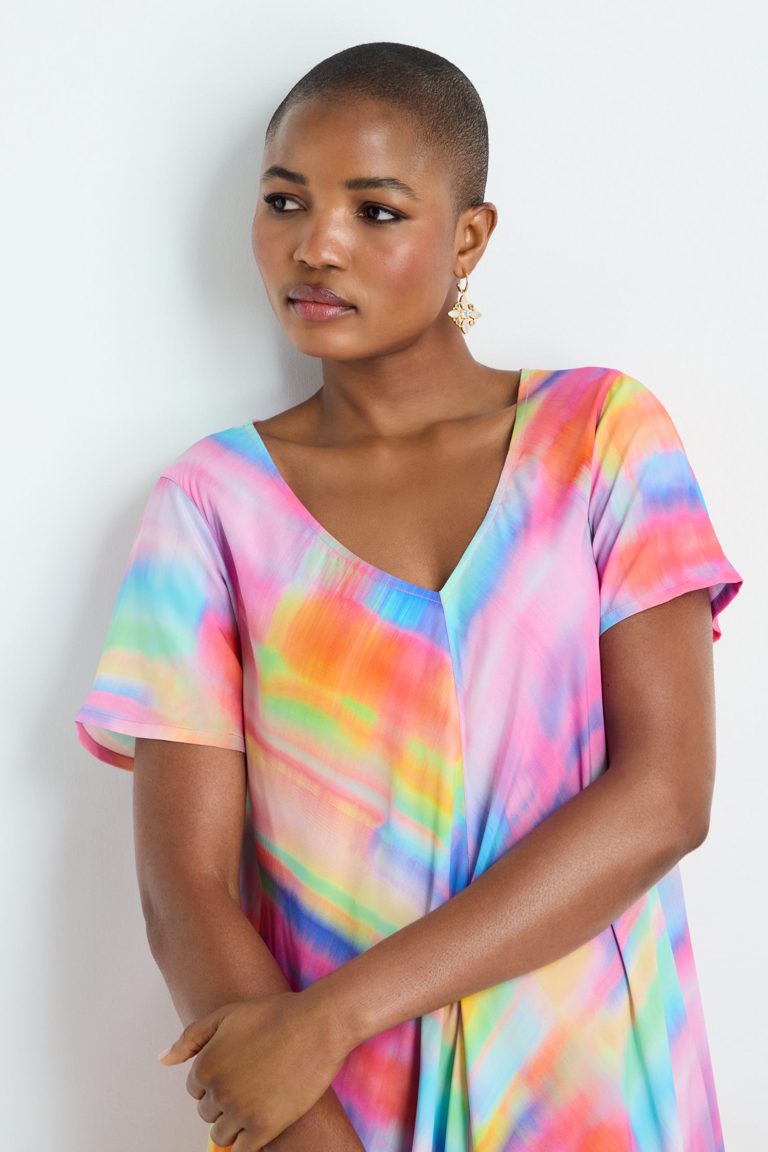 A person with short hair models the Painted Horizon Bubble Dress, featuring a colorful tie-dye design, while standing against a white background and looking slightly to the side.