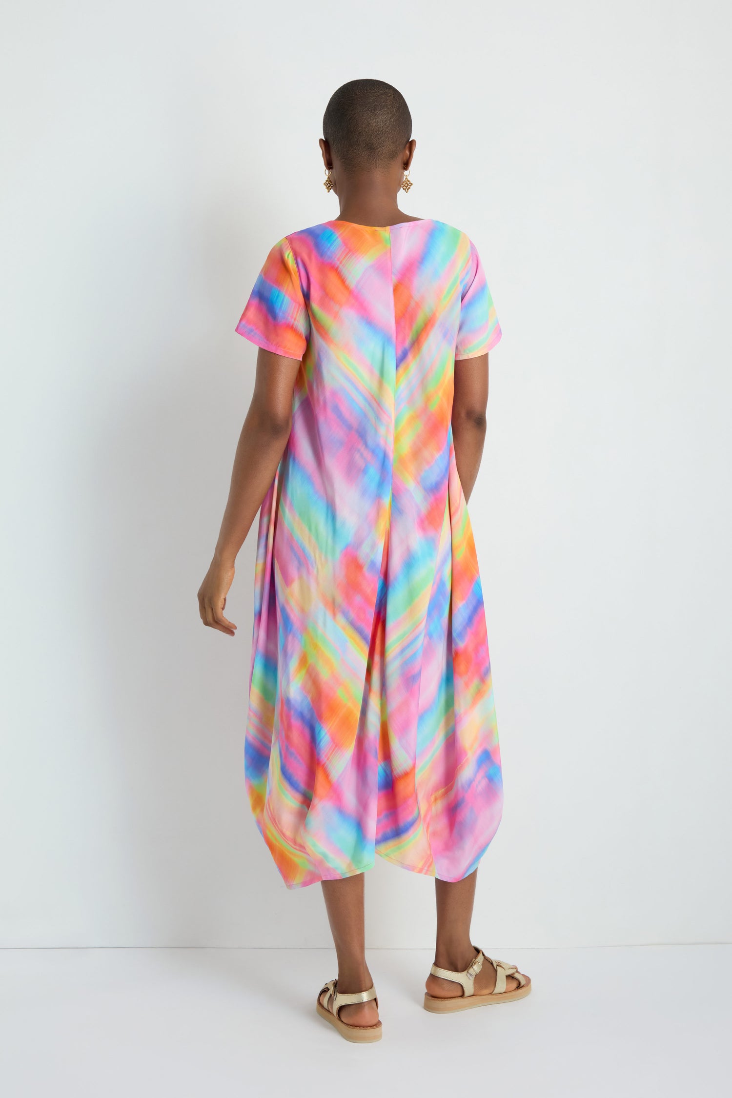 A person stands against a white wall, facing away from the camera, wearing the Painted Horizon Bubble Dress—loose-fitting, bias cut, multicolored with short sleeves—and sandals.