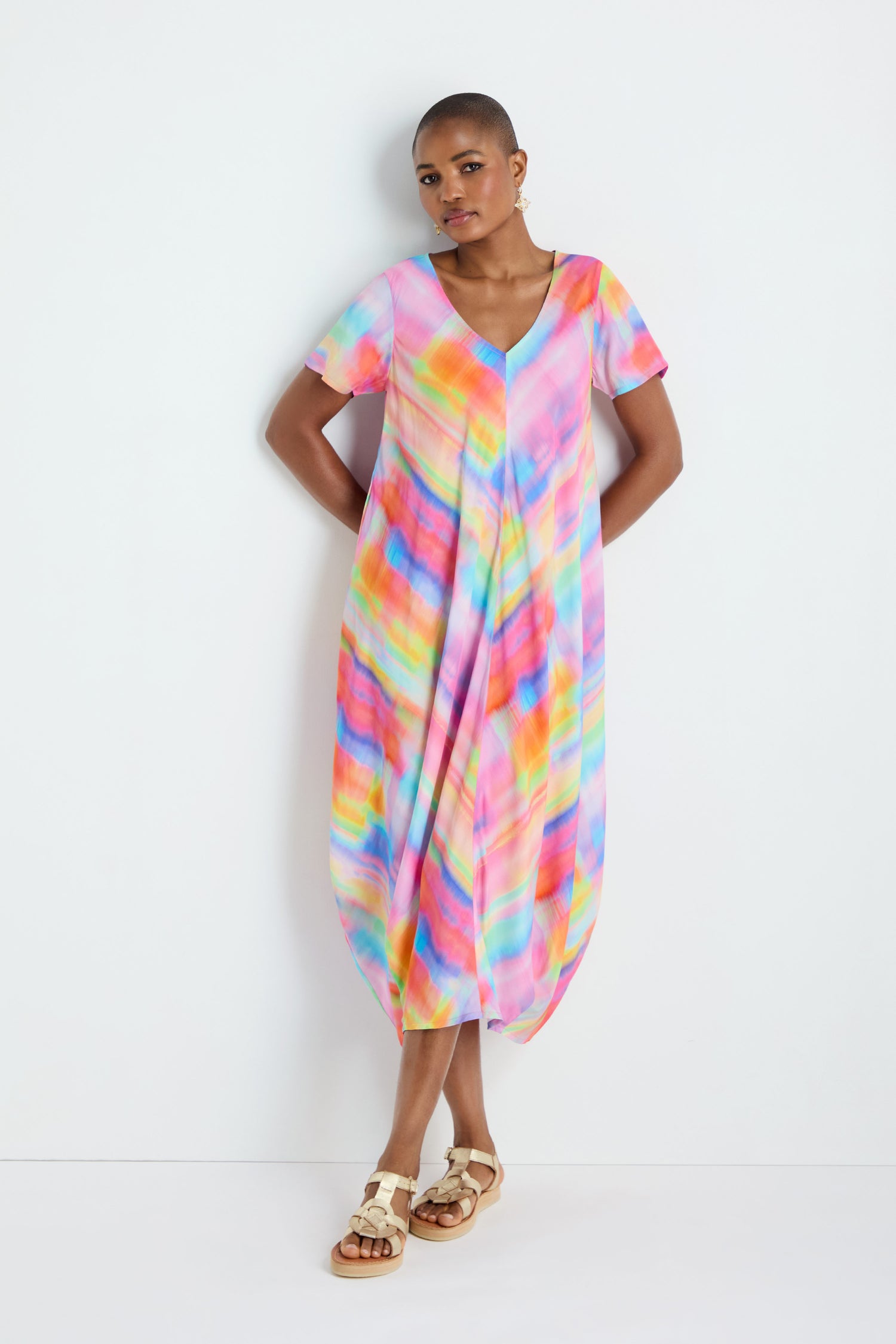 A person wearing the Painted Horizon Bubble Dress, a loose multicolored tie-dye outfit, stands against a white wall with arms relaxed by their sides and beige sandals on.