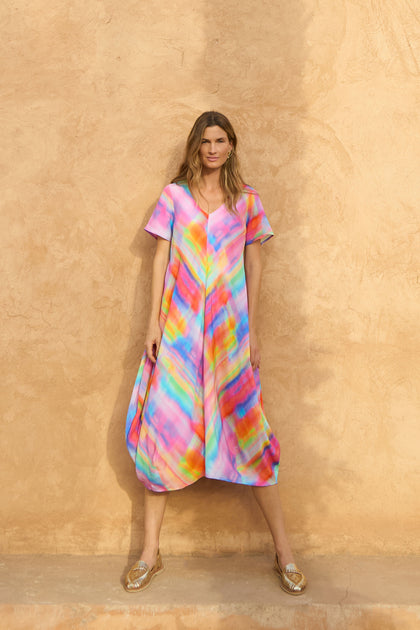 A woman stands against a textured beige wall, wearing the Painted Horizon Bubble Dress and gold flat sandals.