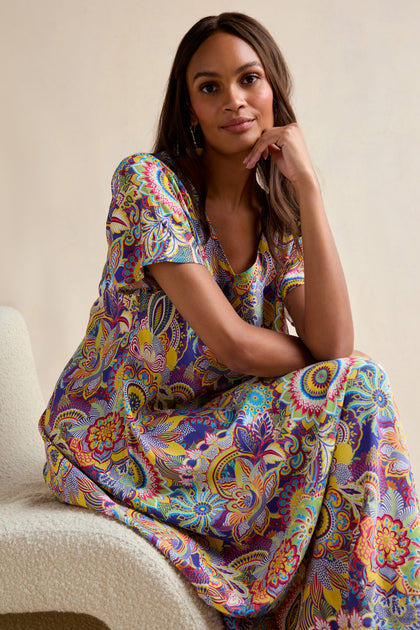 A woman sits on a chair, wearing the Summer Sarasa Bubble Dress, featuring a vibrant floral and paisley print in silk-like viscose, and gazes at the camera with a relaxed expression.