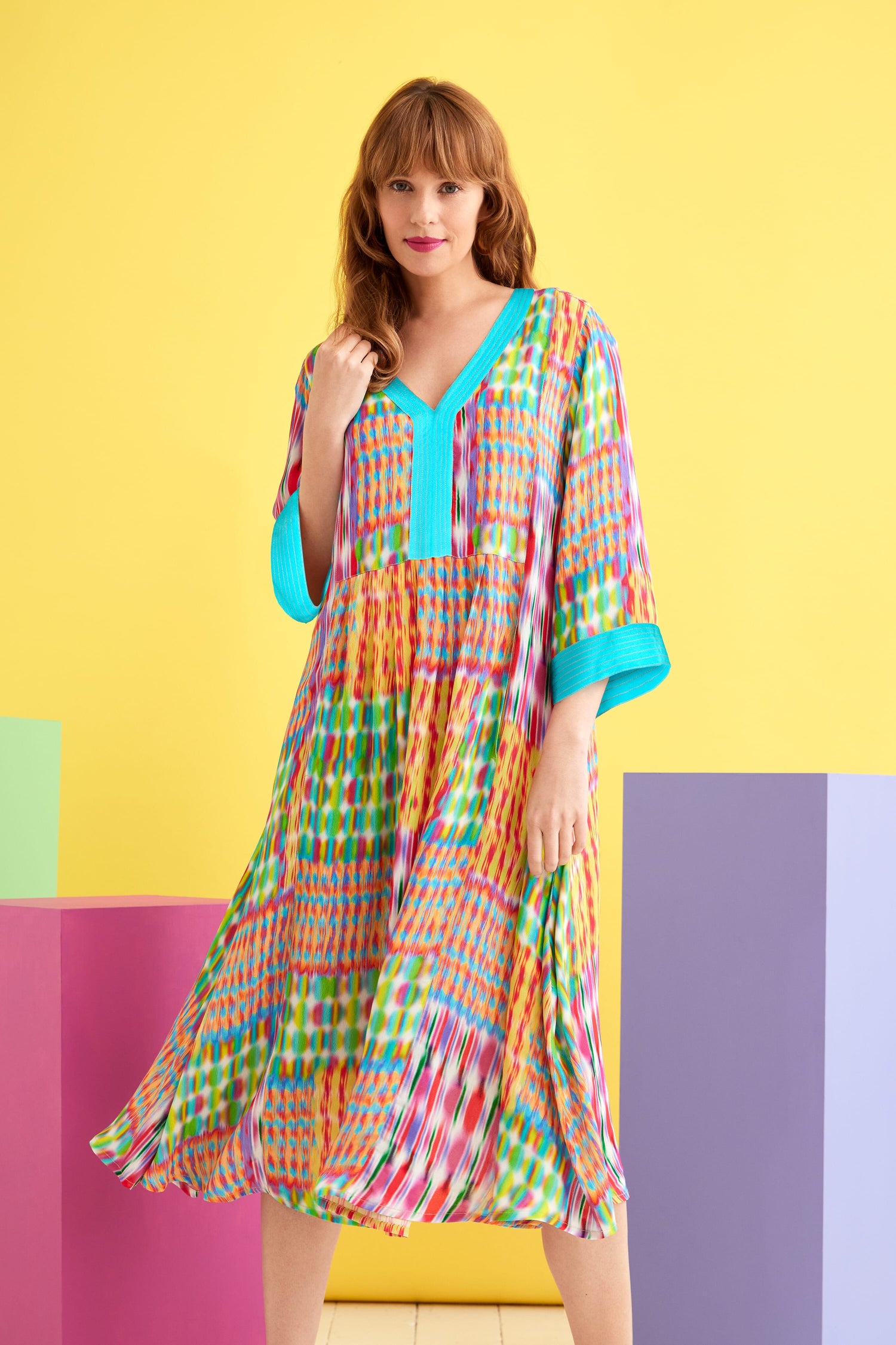 A woman in a Kaleidoscope Ikat Flare Dress with an embroidered turquoise neckline standing in front of colorful cubes.