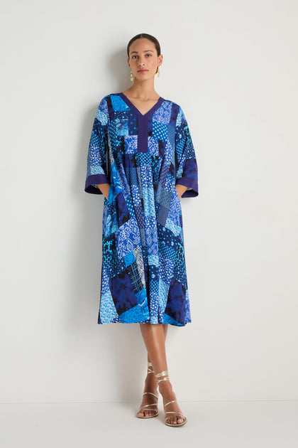 A woman stands against a white wall wearing the Sahara Indigo Patchwork Flared Dress in viscose crepe, featuring a loose, flared silhouette with short sleeves, paired with gold strappy sandals.