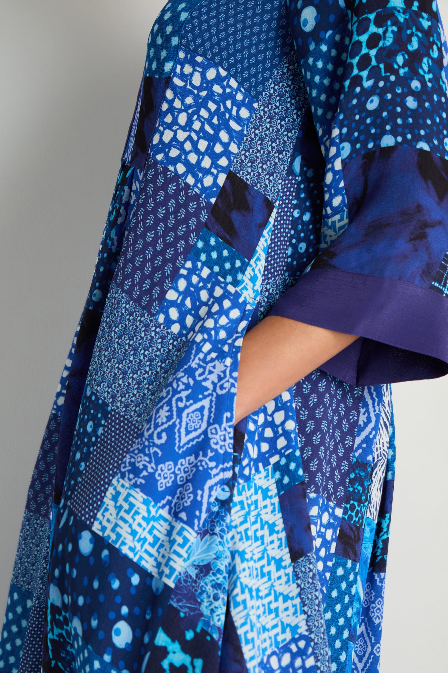 Someone wears the Sahara Indigo Patchwork Flared Dress, made from viscose crepe with blue geometric and abstract patchwork patterns, their hand tucked into a side pocket.