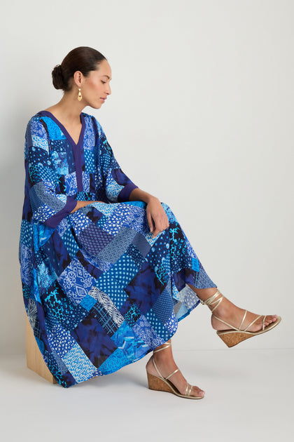 A woman sits sideways on a wooden block against a white background, wearing the Sahara Indigo Patchwork Flared Dress paired with gold wedge sandals and drop earrings.