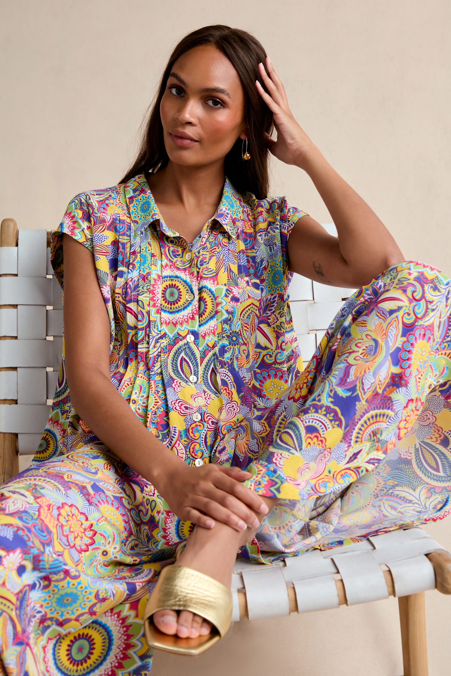 A woman wearing the oversized, colorful Summer Sarasa Jumpsuit sits on a woven chair, resting one hand on her head and looking at the camera.