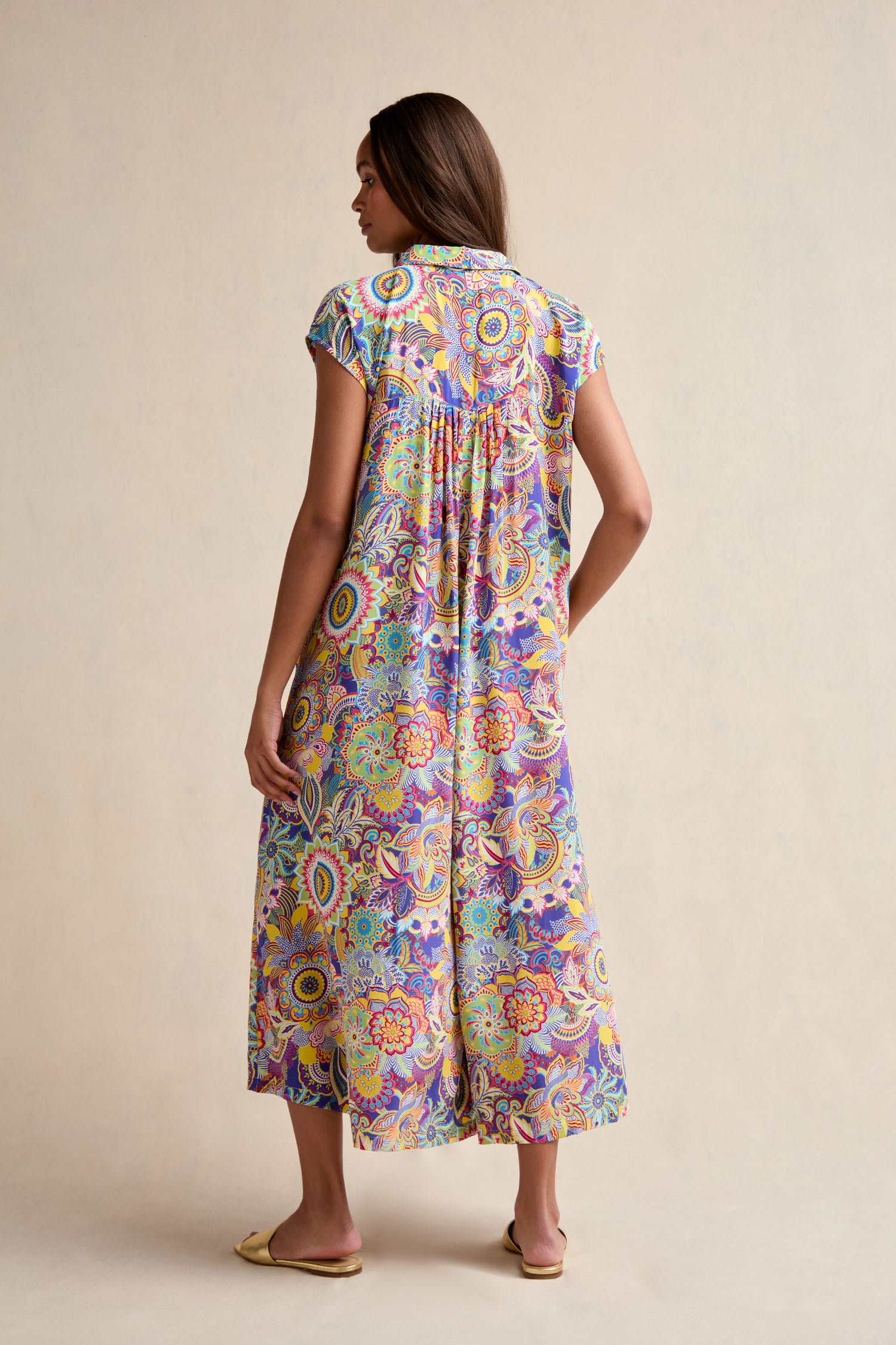 A woman with long hair faces away, wearing the Summer Sarasa Jumpsuit—a colorful, short-sleeved one-piece featuring vibrant paisley and floral patterns. She pairs it with flat shoes.