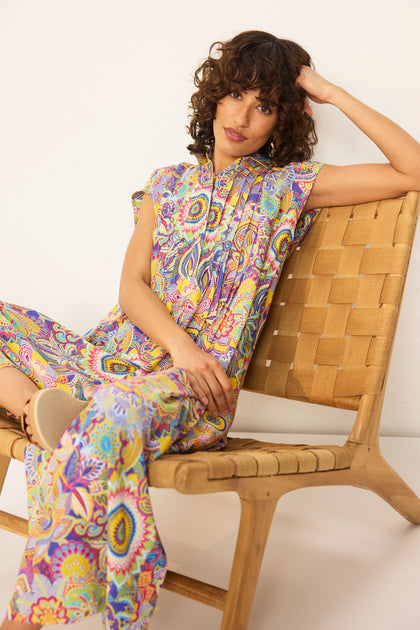 A woman with curly hair sits on a woven chair, wearing the Summer Sarasa Jumpsuit—short-sleeved, wide-leg pants in colorful patterned silk-like viscose—against a plain white background.