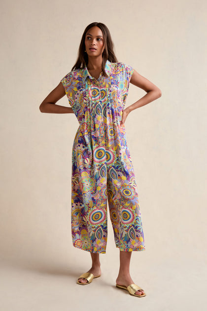 A woman stands with hands on hips, wearing the Summer Sarasa Jumpsuit—featuring wide legs and vibrant patterns—paired with yellow sandals against a plain beige background.