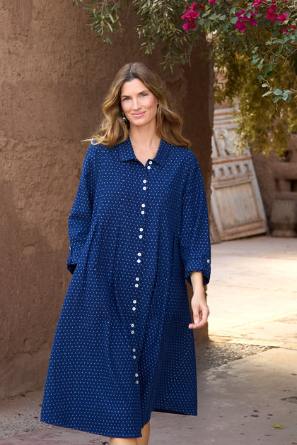 A woman wearing the Indigo Spot Cotton Shirt Dress, featuring blue cotton fabric, long sleeves, a spot pattern, and an A-line silhouette, walks outdoors along a sunlit adobe wall with greenery above.