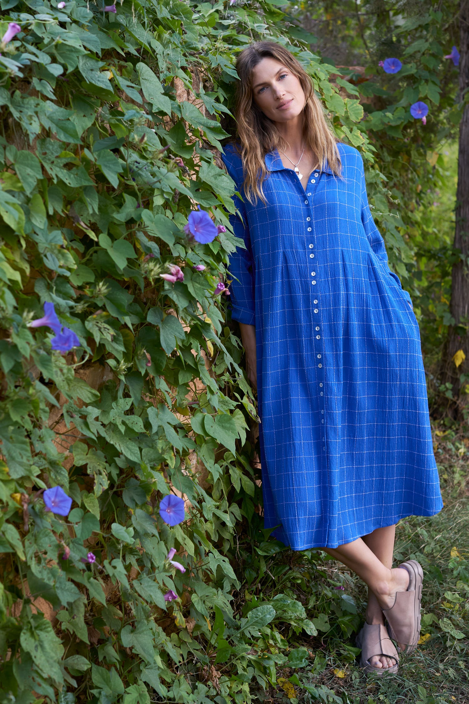 A woman in a Stitched Grid Check Dress displays a flattering silhouette while leaning against a vine-covered wall with purple flowers, exuding timeless style as she looks at the camera.