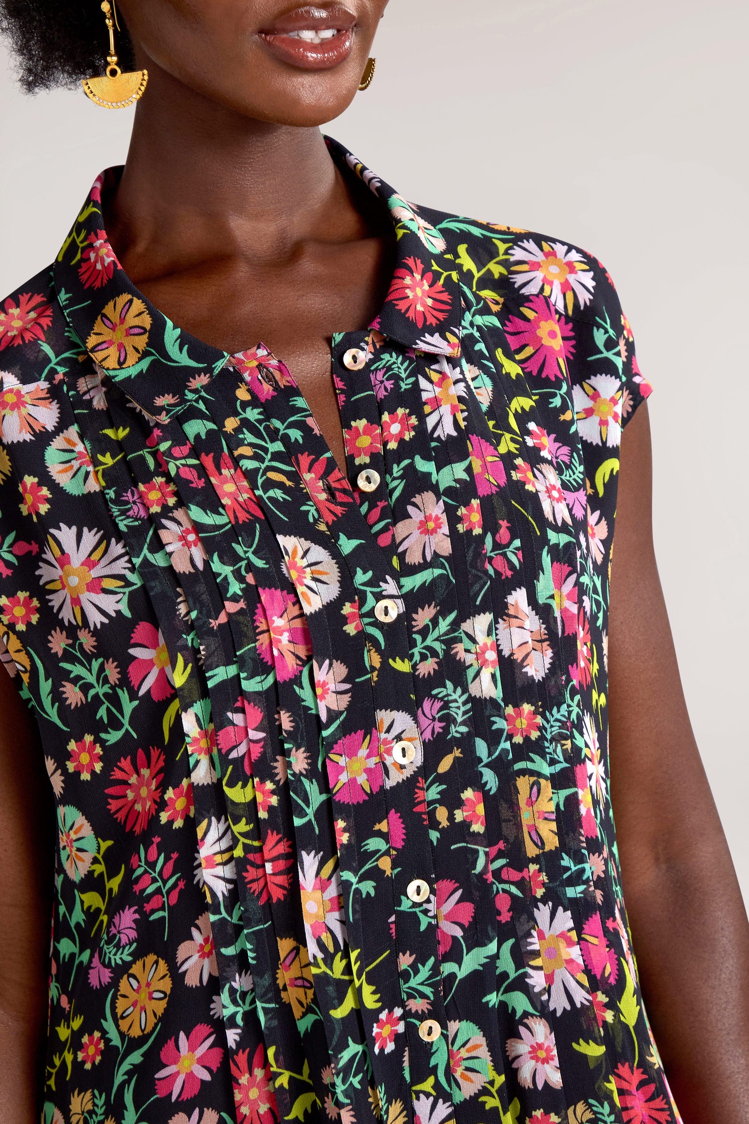 Close-up of a person wearing a Folkloric Floral Shirt Dress and gold earrings against a neutral background. The blouse, with its silk-like feel, has short sleeves and small buttons down the front.