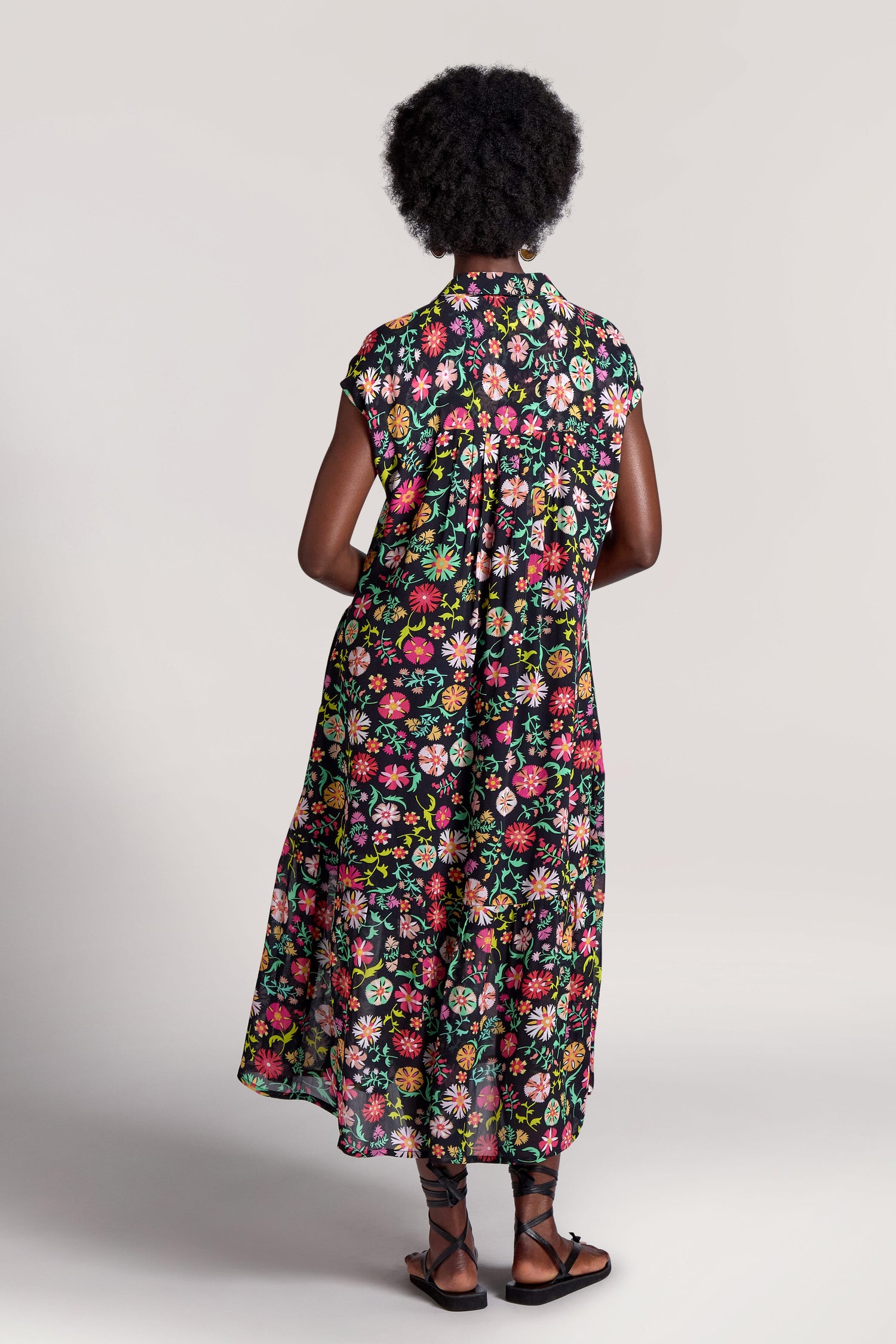 Person with short curly hair, standing with their back facing the camera, wearing a Folkloric Floral Shirt Dress and black sandals against a plain background.