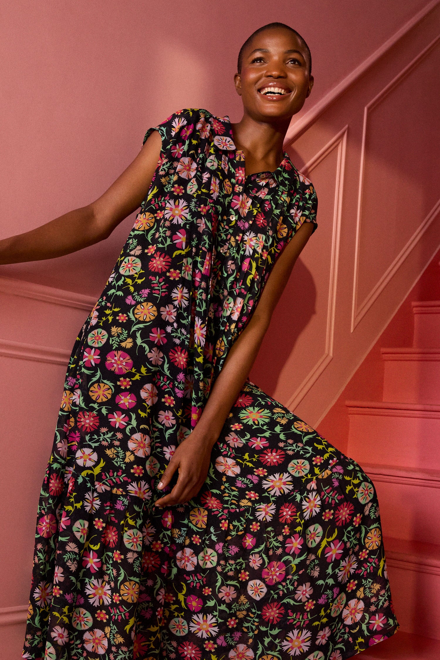 A person wearing a Folkloric Floral Shirt Dress stands in front of a pink wall and staircase, smiling.