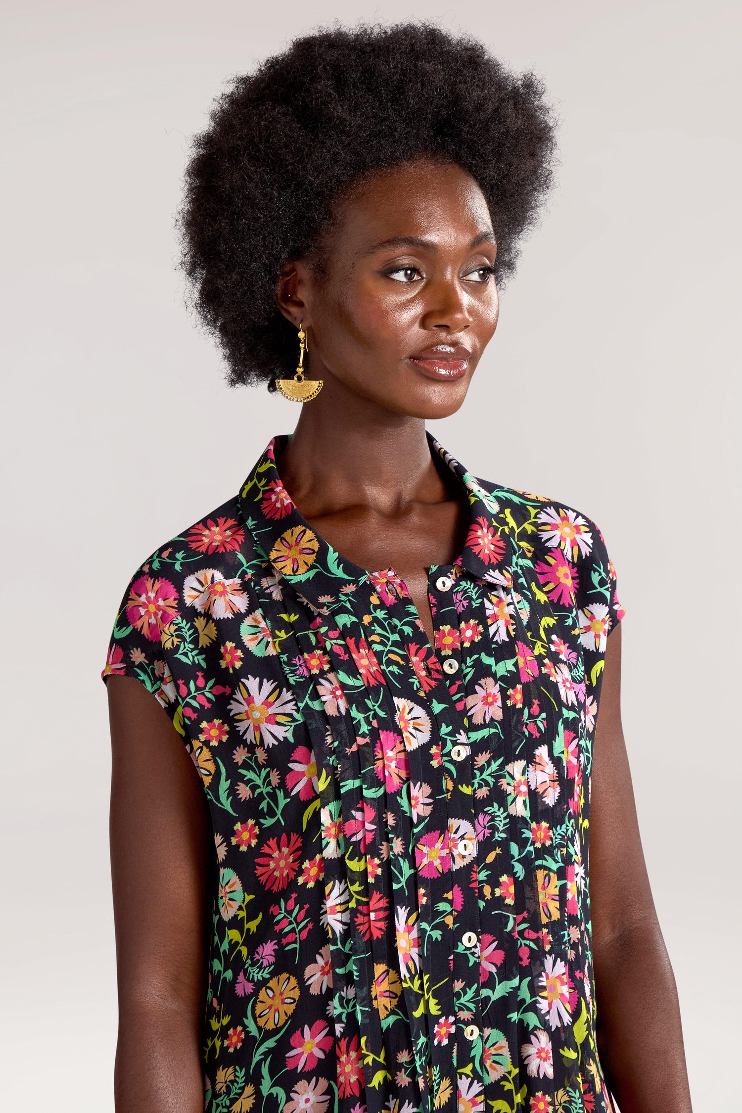 A person with an afro hairstyle wears a short-sleeved, Folkloric Floral Shirt Dress with a silk-like feel and gold earrings, looking slightly to the side.