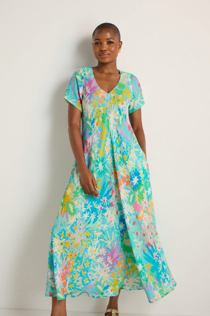 A person stands against a white background wearing the Floral Oasis Maxi Dress, made from soft viscose crepe, paired with sandals.