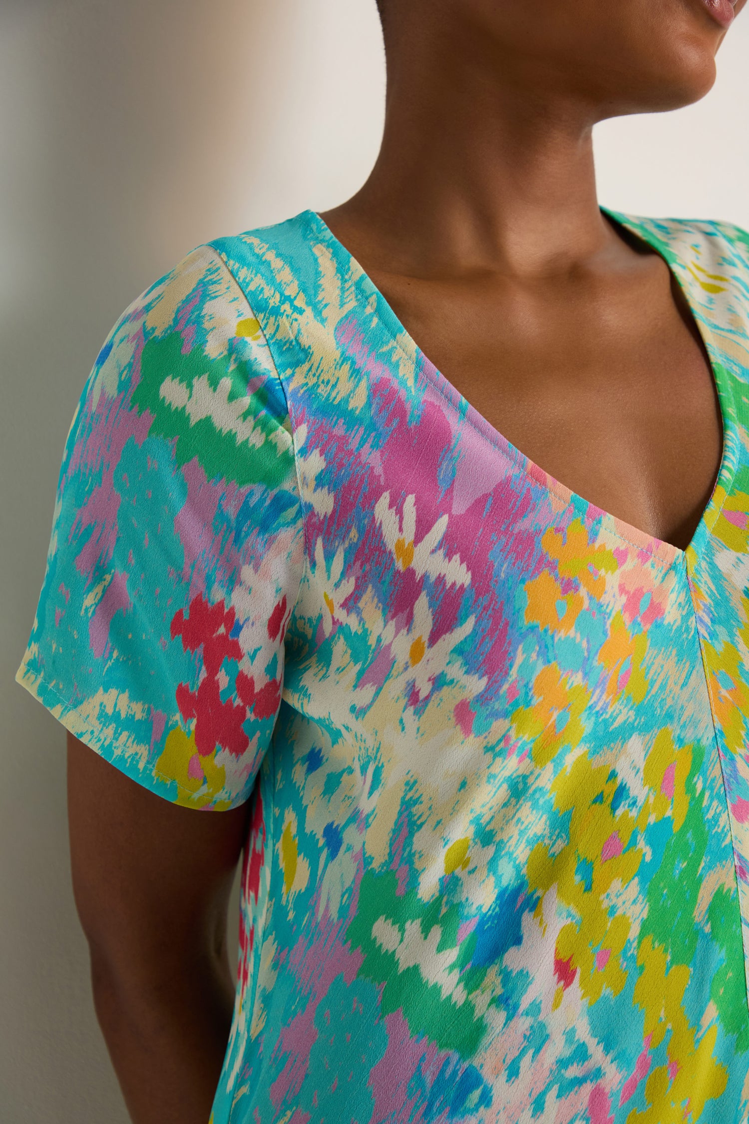 A person wears the Floral Oasis Maxi Dress, a short-sleeved piece in soft viscose crepe with an abstract floral pattern in turquoise, yellow, pink, and white. Only the upper body is visible.