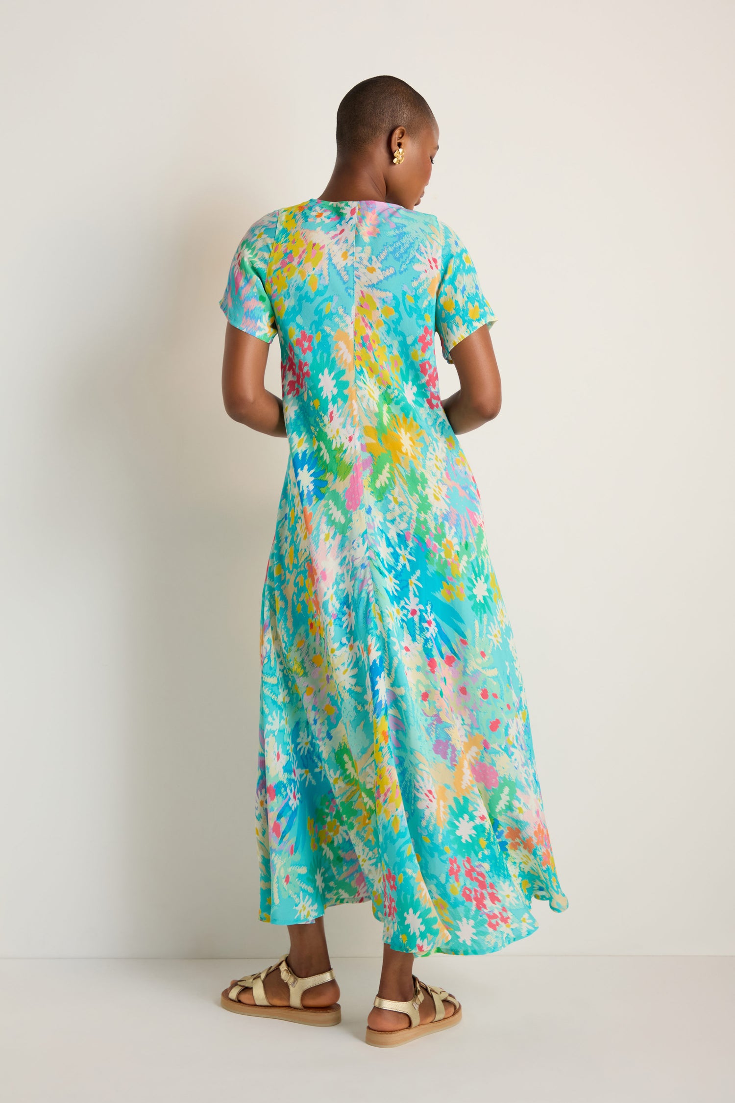 A person stands with their back to the camera wearing the Floral Oasis Maxi Dress in light blue and tan sandals, set against a plain light background.