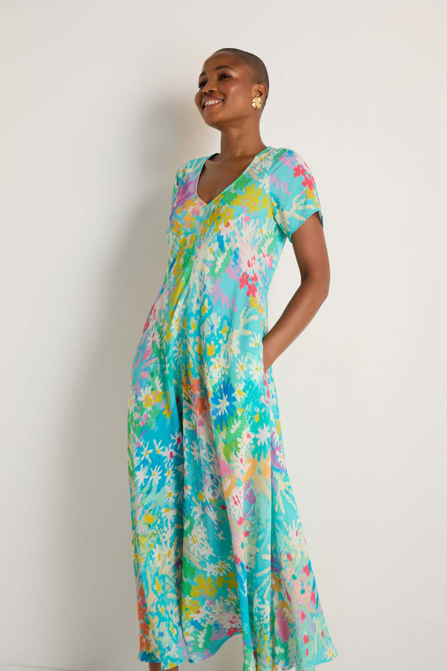 A woman wearing the Floral Oasis Maxi Dress, featuring a soft viscose crepe fabric with a vibrant floral print and short sleeves, stands against a plain white background, smiling as she looks to the side.