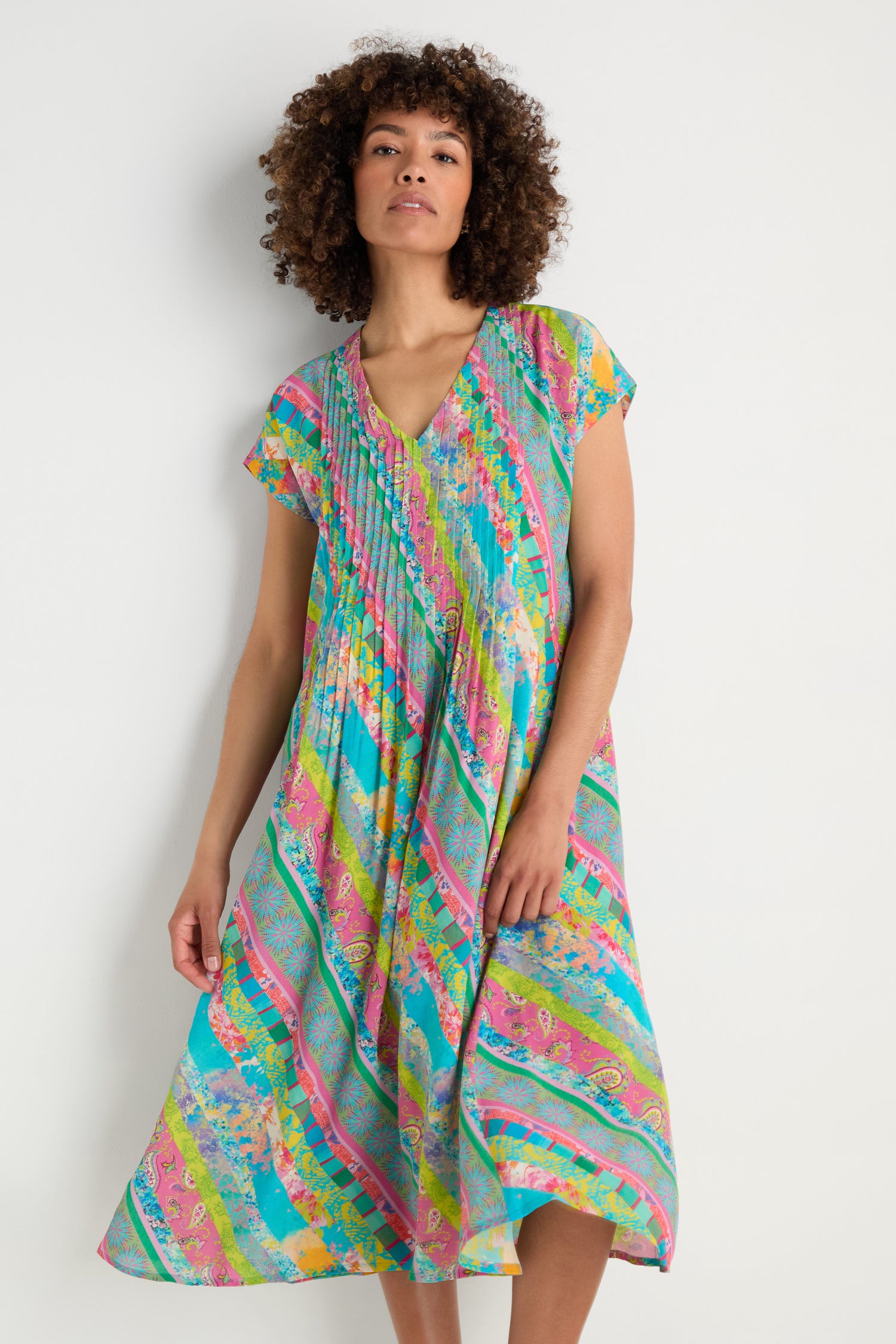 A woman stands against a plain background wearing the Patchwork Stripe Pleated Dress, featuring colorful patterns, short sleeves, a V-neck, pleats, and a flattering fit-and-flare silhouette.