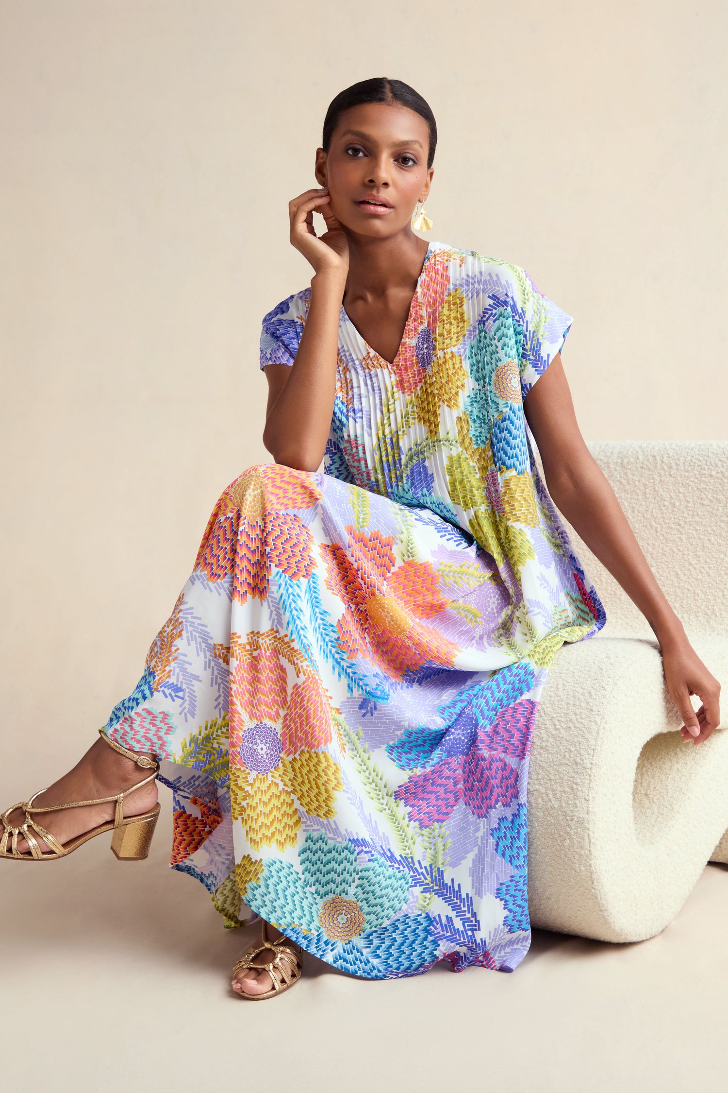 A woman sits on a curved white chair against a neutral background, wearing the Pixelated Petals Print Dress with short sleeves and gold heeled sandals.