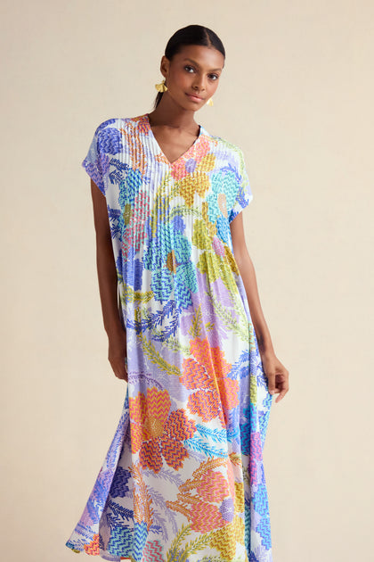 A woman stands against a plain light background wearing the Pixelated Petals Print Dress—loose-fitting with short sleeves, a V-neck, and a vibrant pixelated floral pattern.
