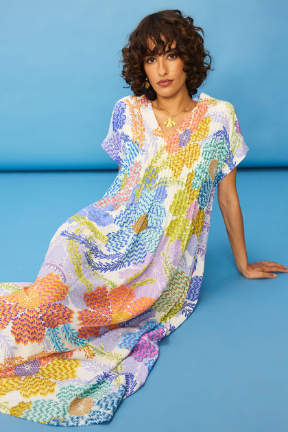 A person with curly hair sits on the floor against a blue background, wearing the loose-fitting Pixelated Petals Print Dress and a gold necklace.