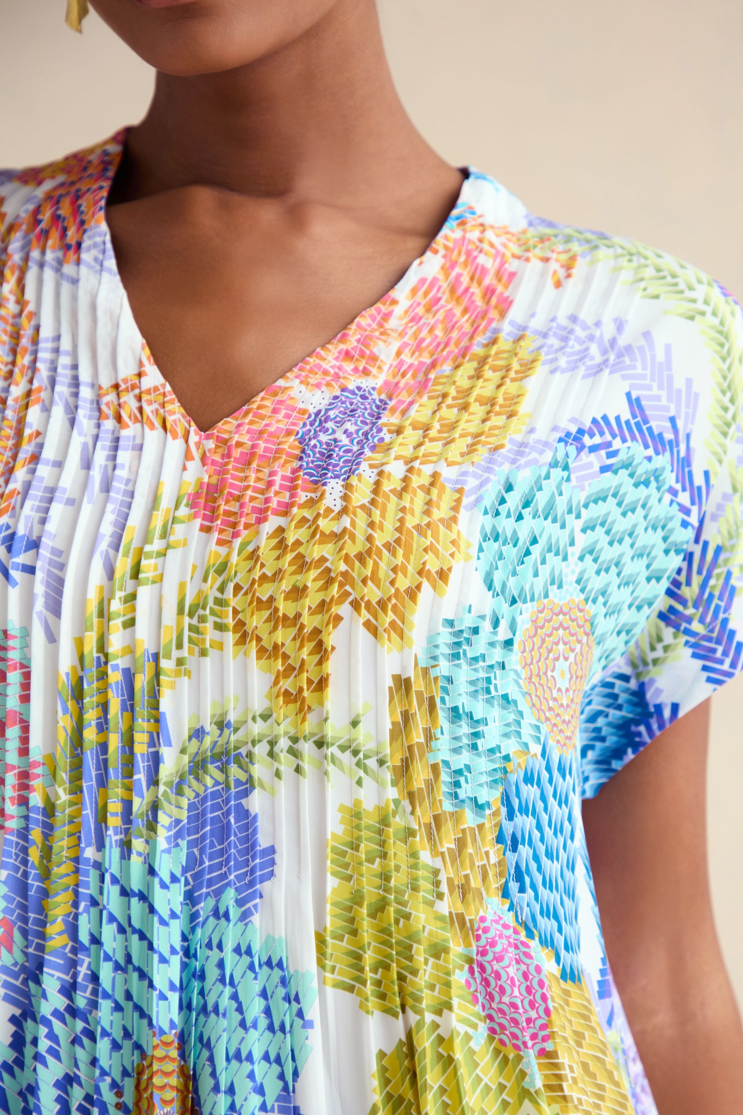 A close-up of a person wearing the Pixelated Petals Print Dress, a pleated V-neck dress with a colorful pixelated floral pattern in blue, yellow, purple, orange, and green.
