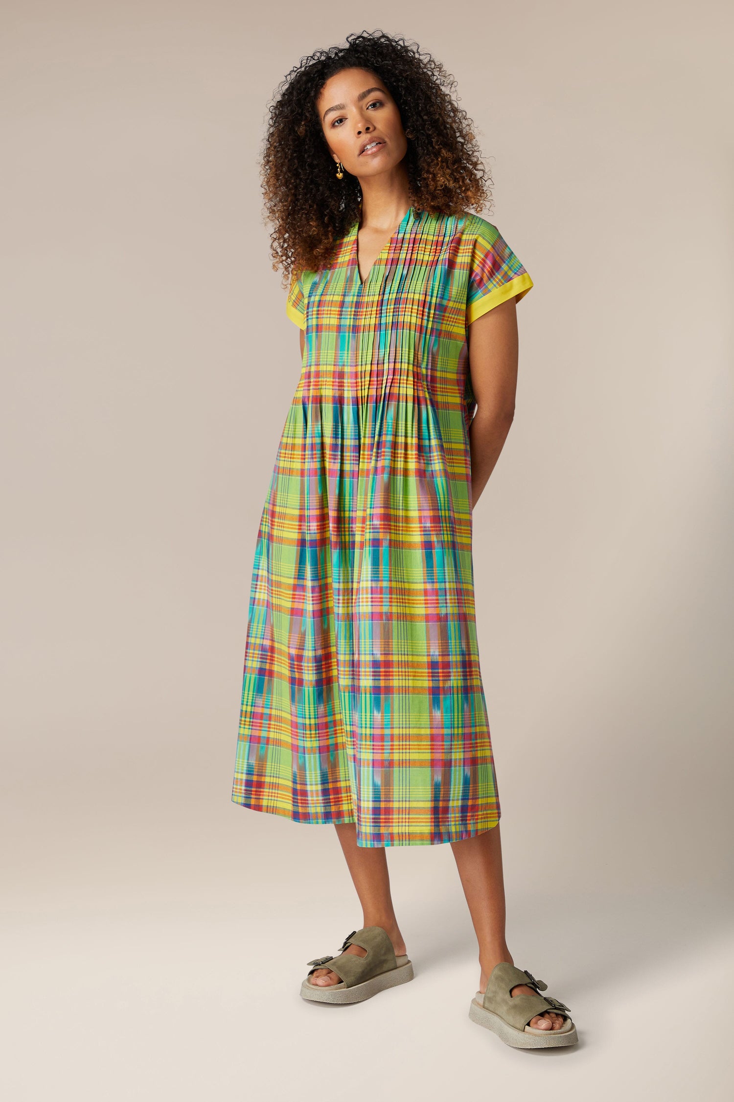A woman with curly hair wearing a Multicoloured Ikat Check Dress and green sandals stands against a neutral background.