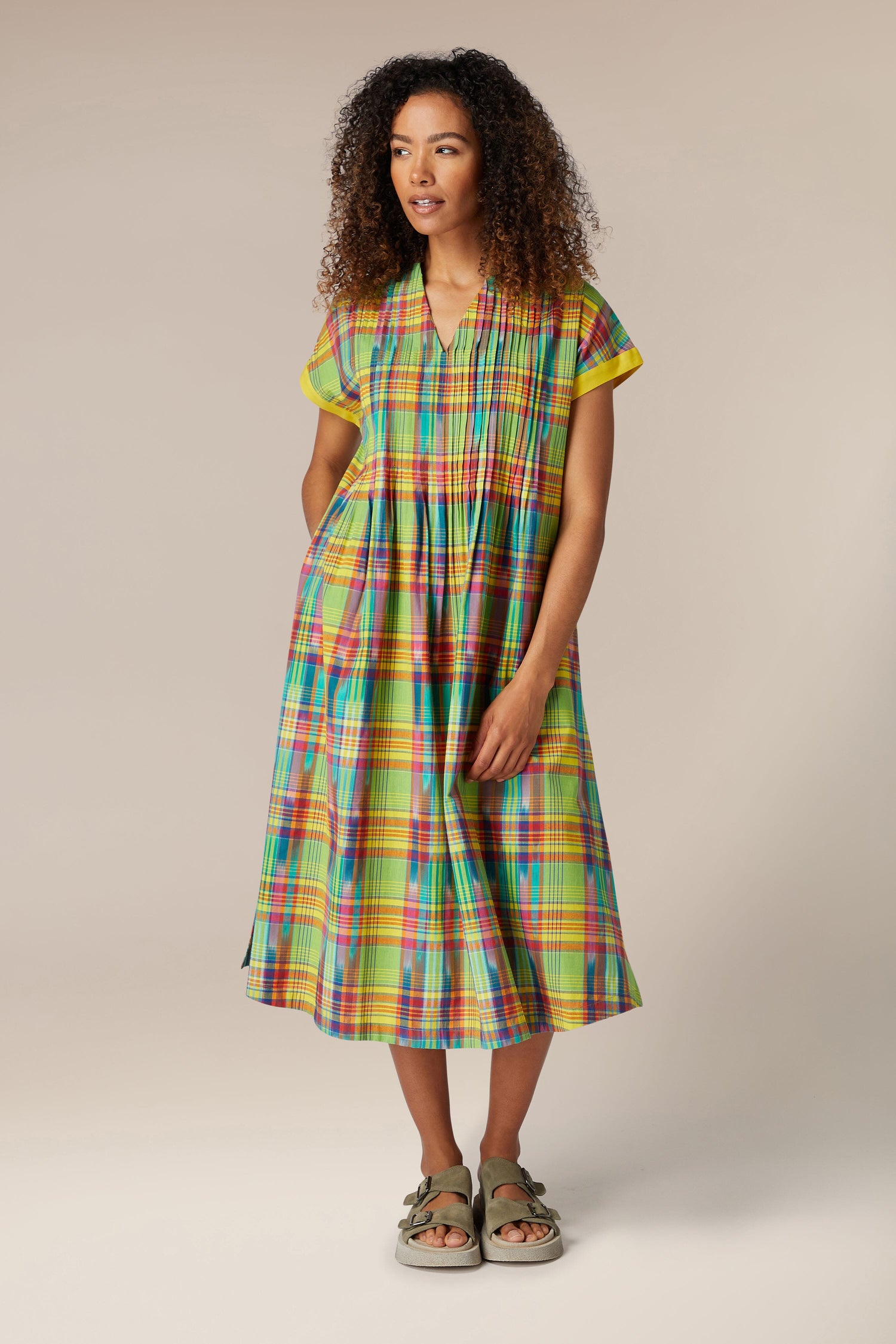 A woman with curly hair wearing a Multicoloured Ikat Check Dress and sandals, standing against a neutral background.