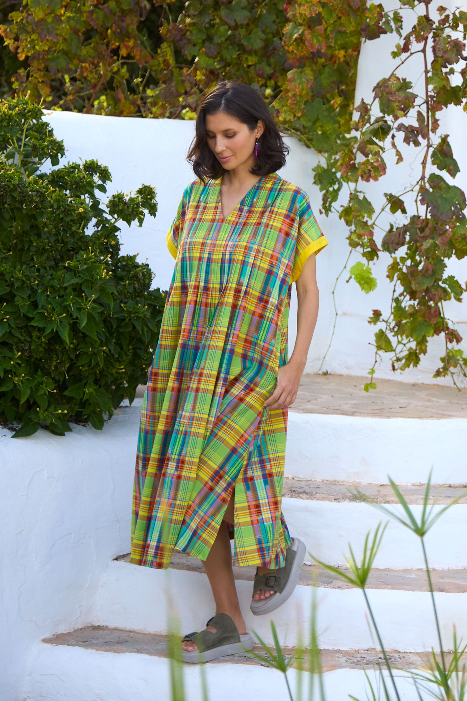 A woman wearing a Multicoloured Ikat Check Dress walks down white outdoor stairs, surrounded by greenery.