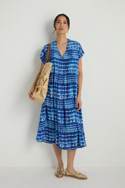 Woman wearing a Tie Dye Printed Viscose Pleated Dress, gold sandals, and carrying a large woven bag, standing against a plain white background.