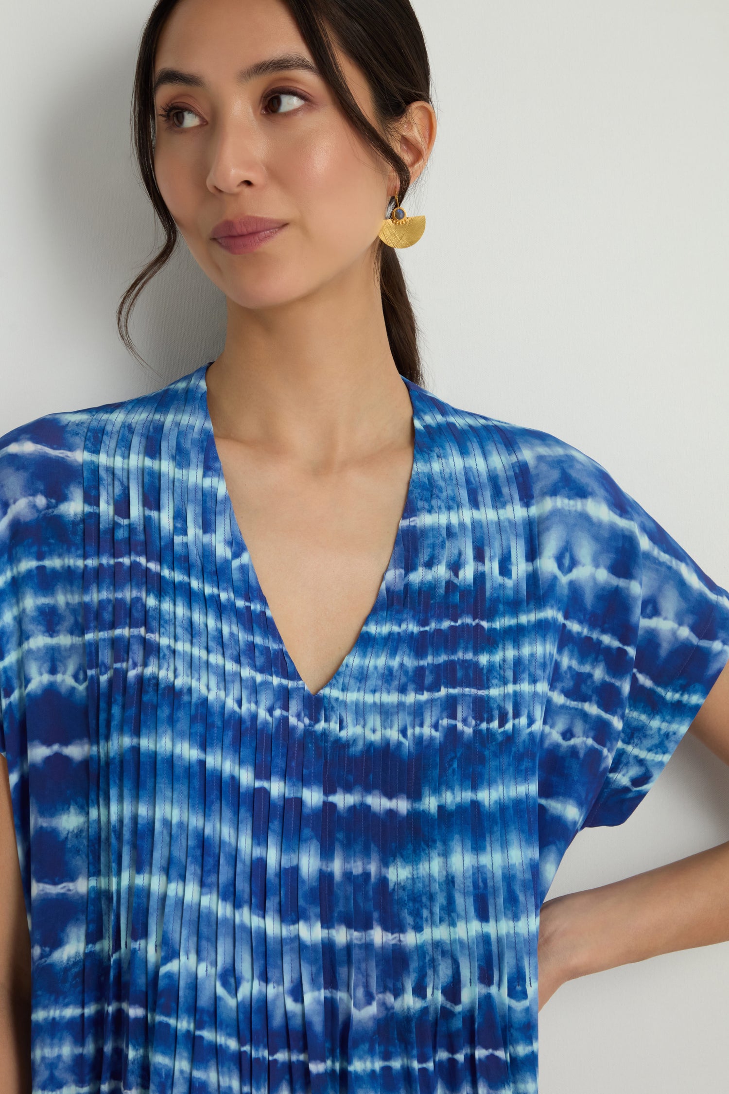 A woman stands against a plain white background wearing the Tie Dye Printed Viscose Pleated Dress and a gold statement earring.
