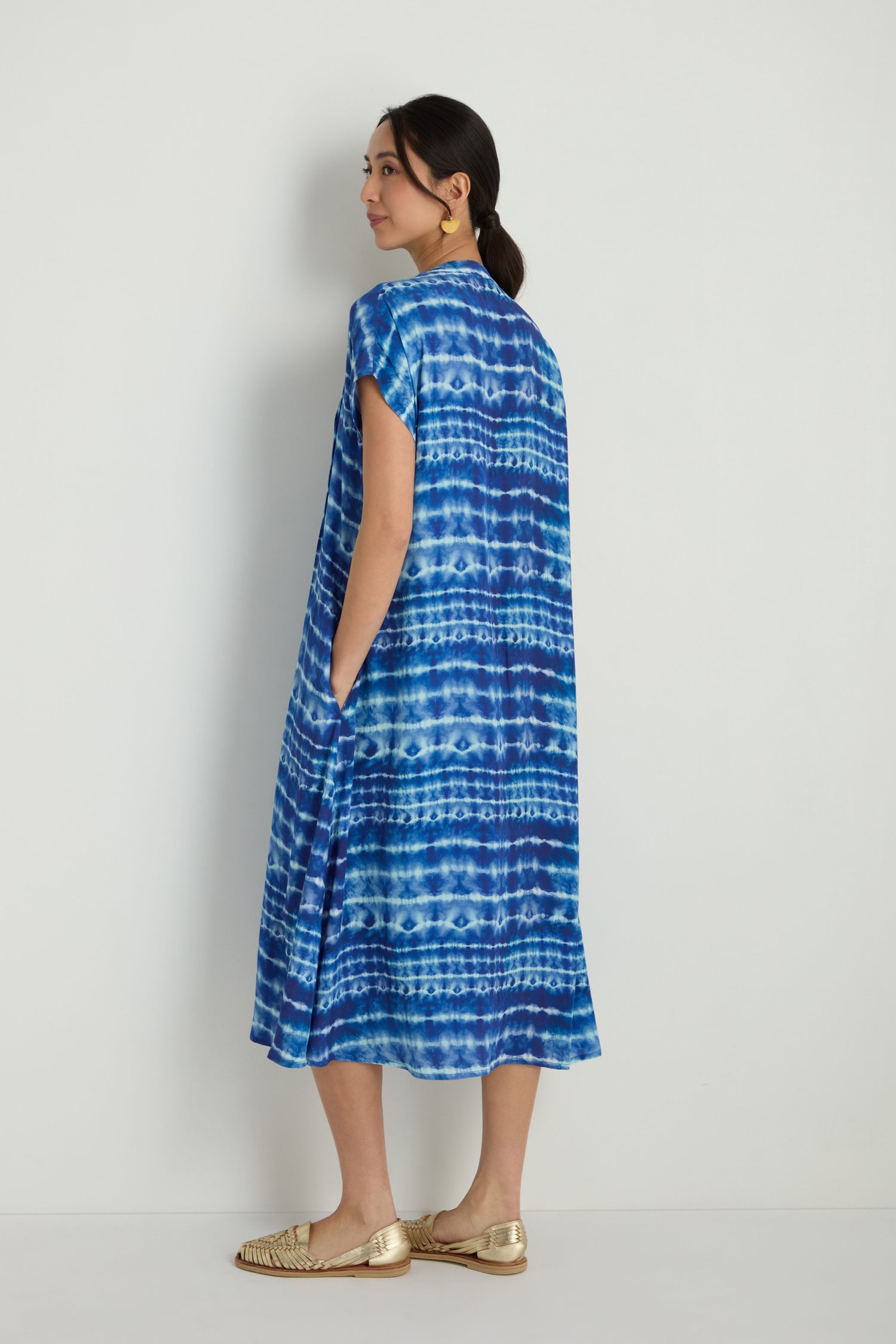 A woman stands sideways against a white wall, wearing the Tie Dye Printed Viscose Pleated Dress and gold woven flat shoes.