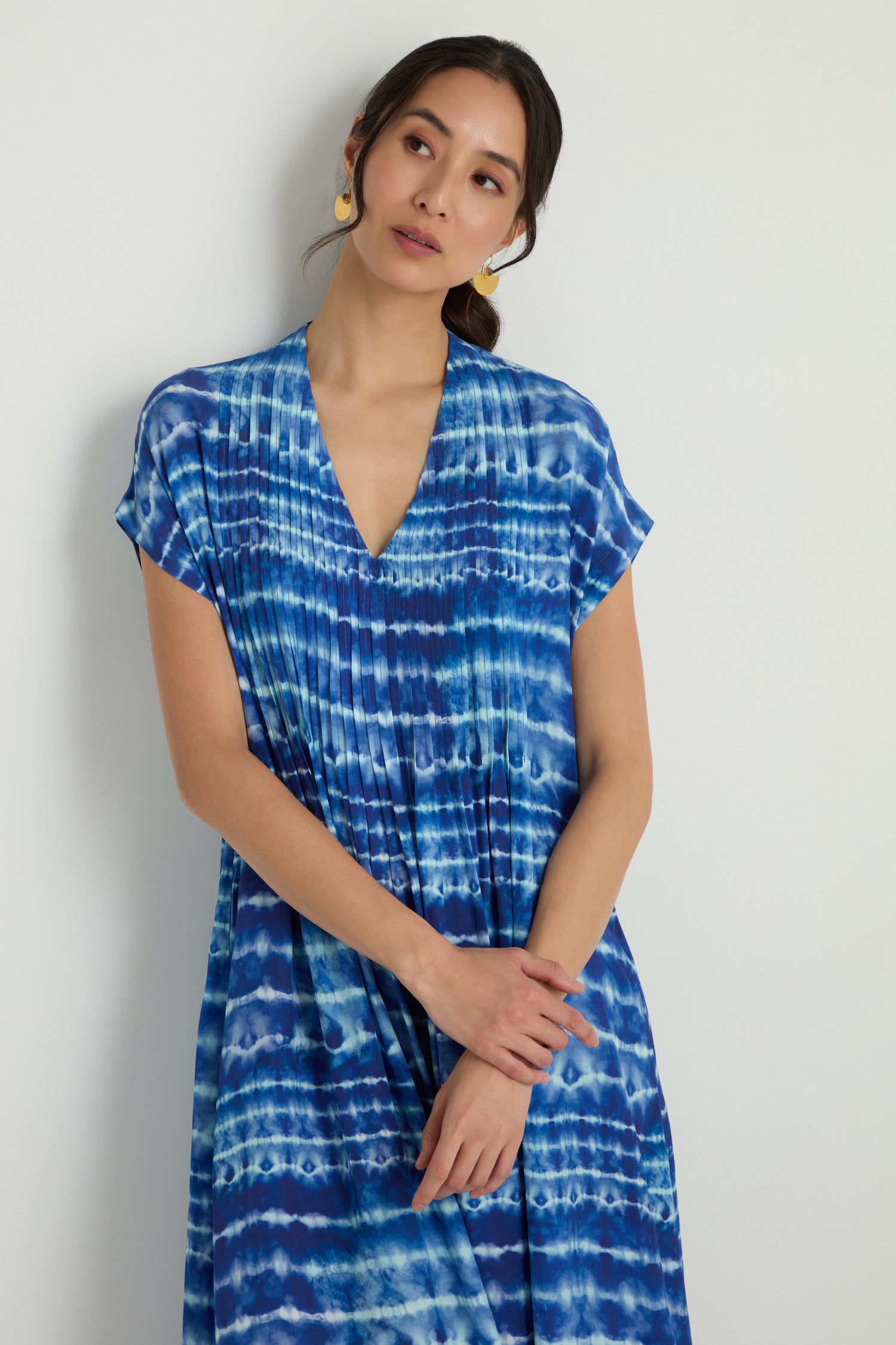 A woman stands against a white wall wearing the Tie Dye Printed Viscose Pleated Dress, featuring short sleeves and a V-neck. The dress combines comfortable viscose material with effortless style in a trendy blue tie dye print.