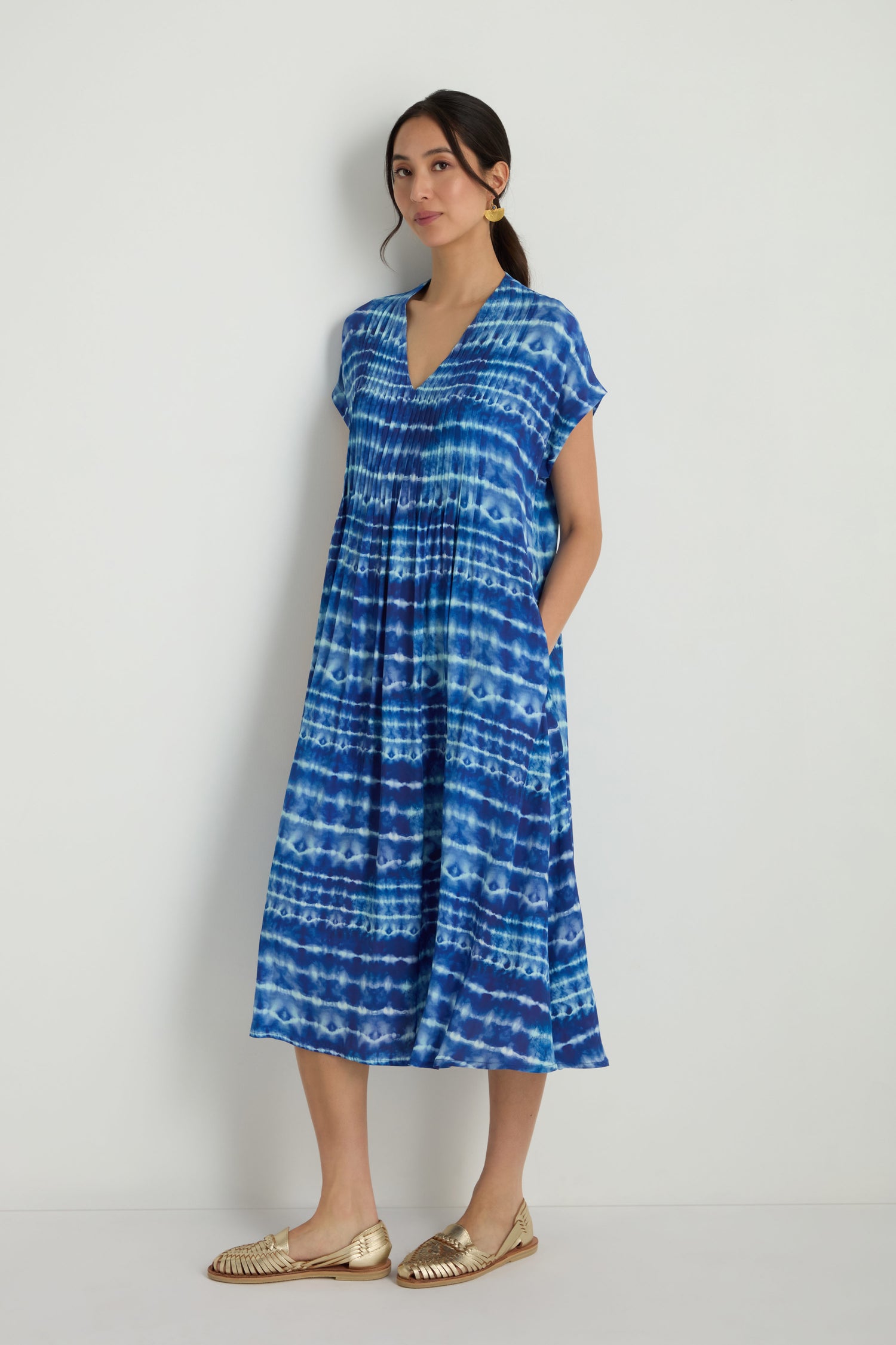 A woman stands against a white wall wearing the Tie Dye Printed Viscose Pleated Dress with short sleeves and gold woven flats.