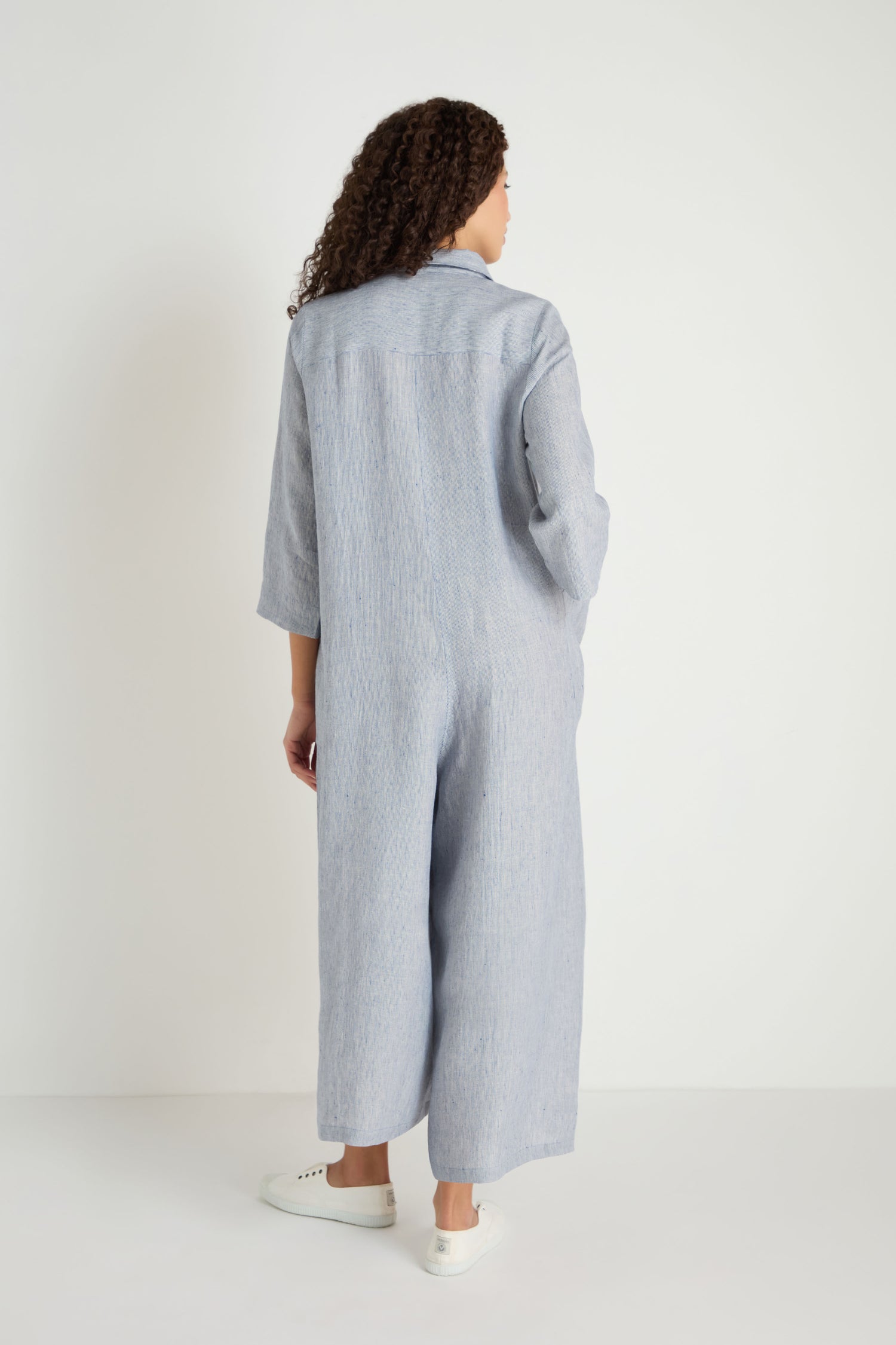 A person with curly hair stands against a white background, facing away, wearing the Ticking Linen Stripe Jumpsuit—a loose-fitting, light blue linen piece with a relaxed silhouette—paired with white sneakers.