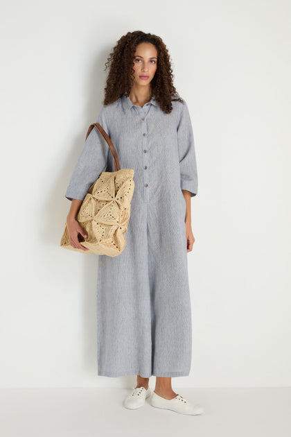 A woman stands against a white background wearing the Ticking Linen Stripe Jumpsuit in light gray, paired with white sneakers and carrying a large woven tote bag on her shoulder.