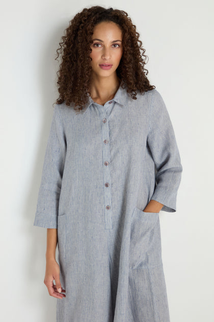 A woman with curly hair wears the Ticking Linen Stripe Jumpsuit, a light blue, long-sleeve button-up style, standing against a plain white background with one hand in her pocket.