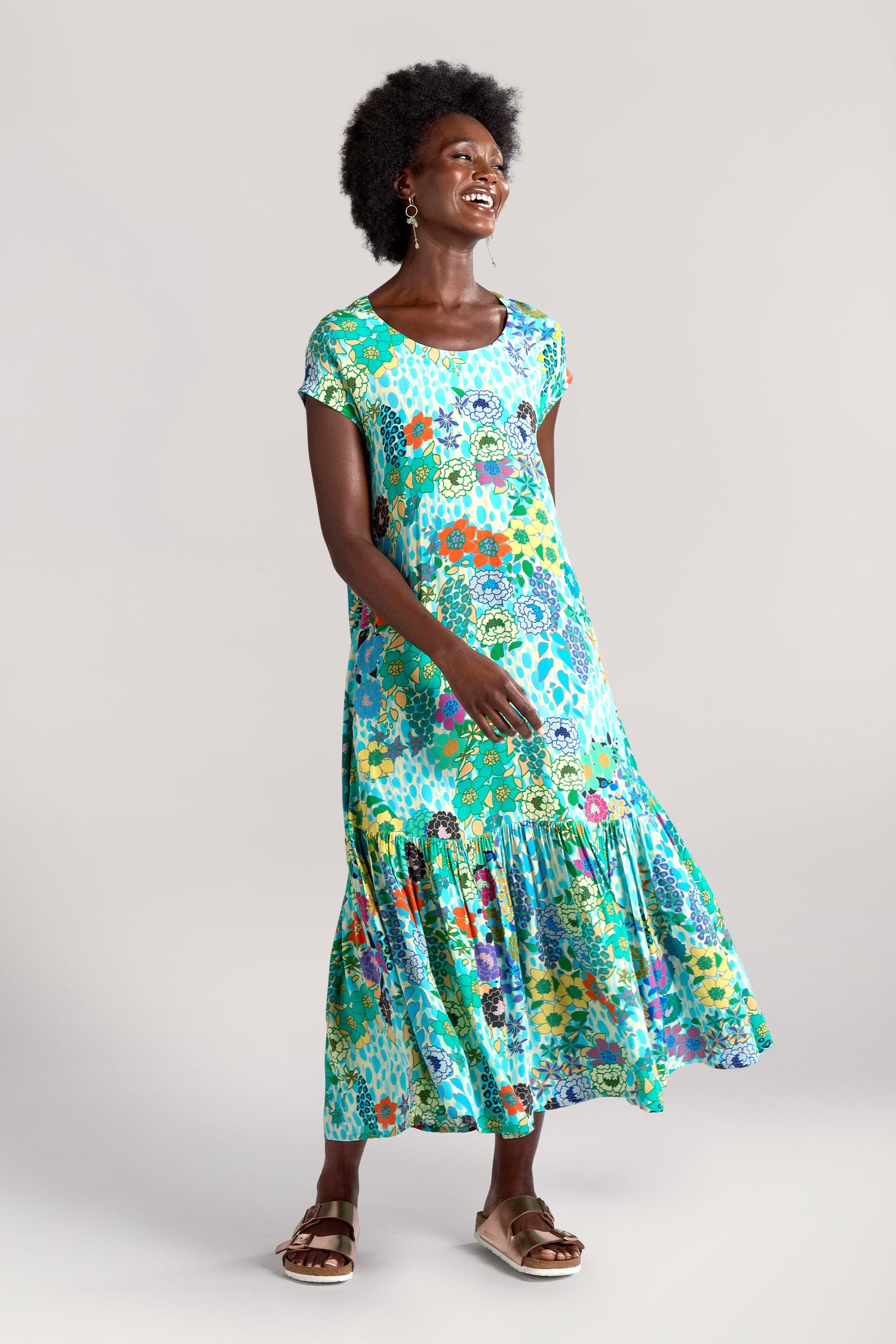 A woman stands smiling, wearing a vibrant Garden Spot Frill Hem Maxi Dress with short sleeves and sandals. She has curly hair and is posing against a plain background.