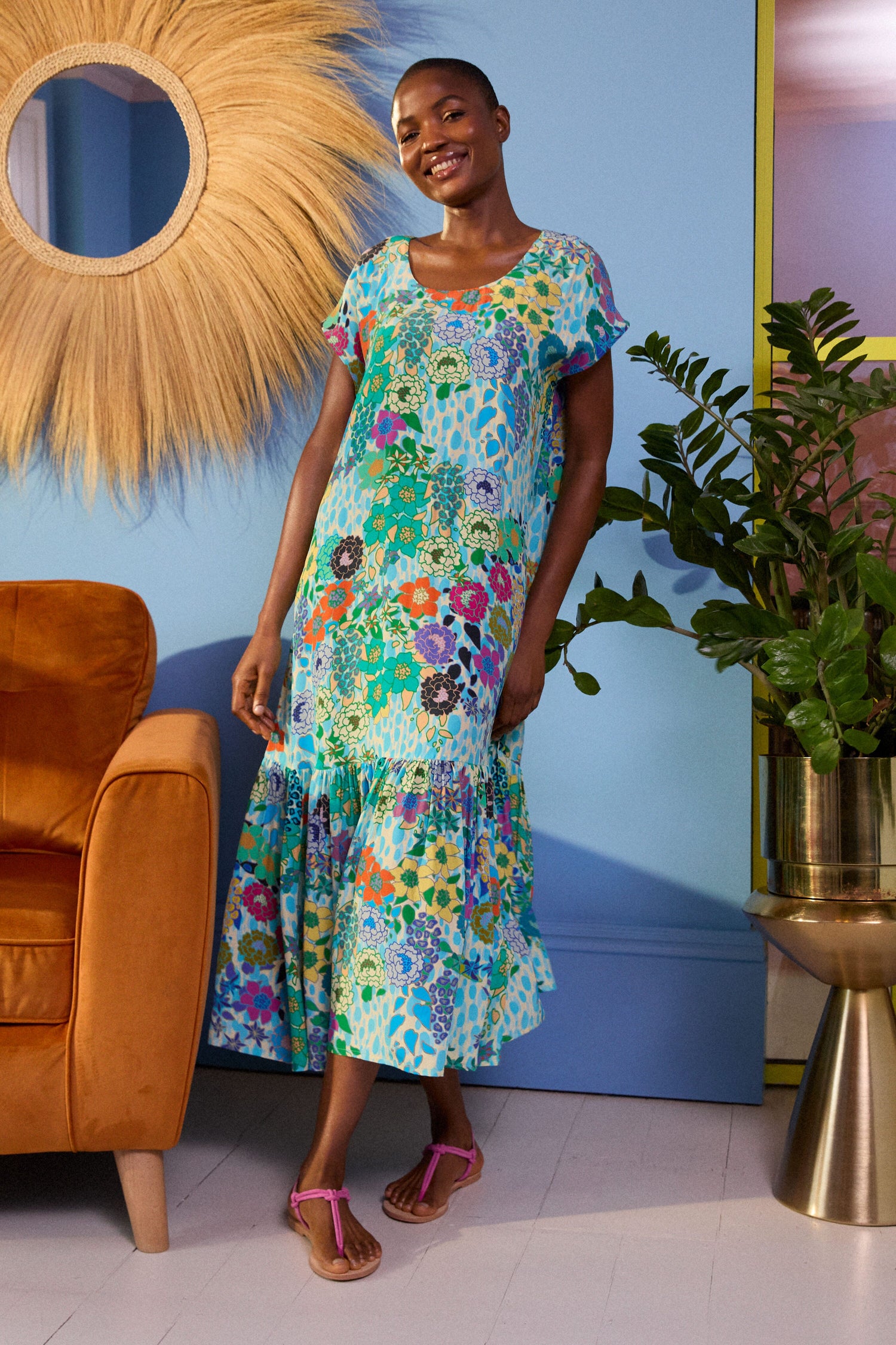 A person wearing a brightly patterned Garden Spot Frill Hem Maxi Dress stands next to a brown chair and a large green plant in a pot, with a decorative wall mirror in the background.