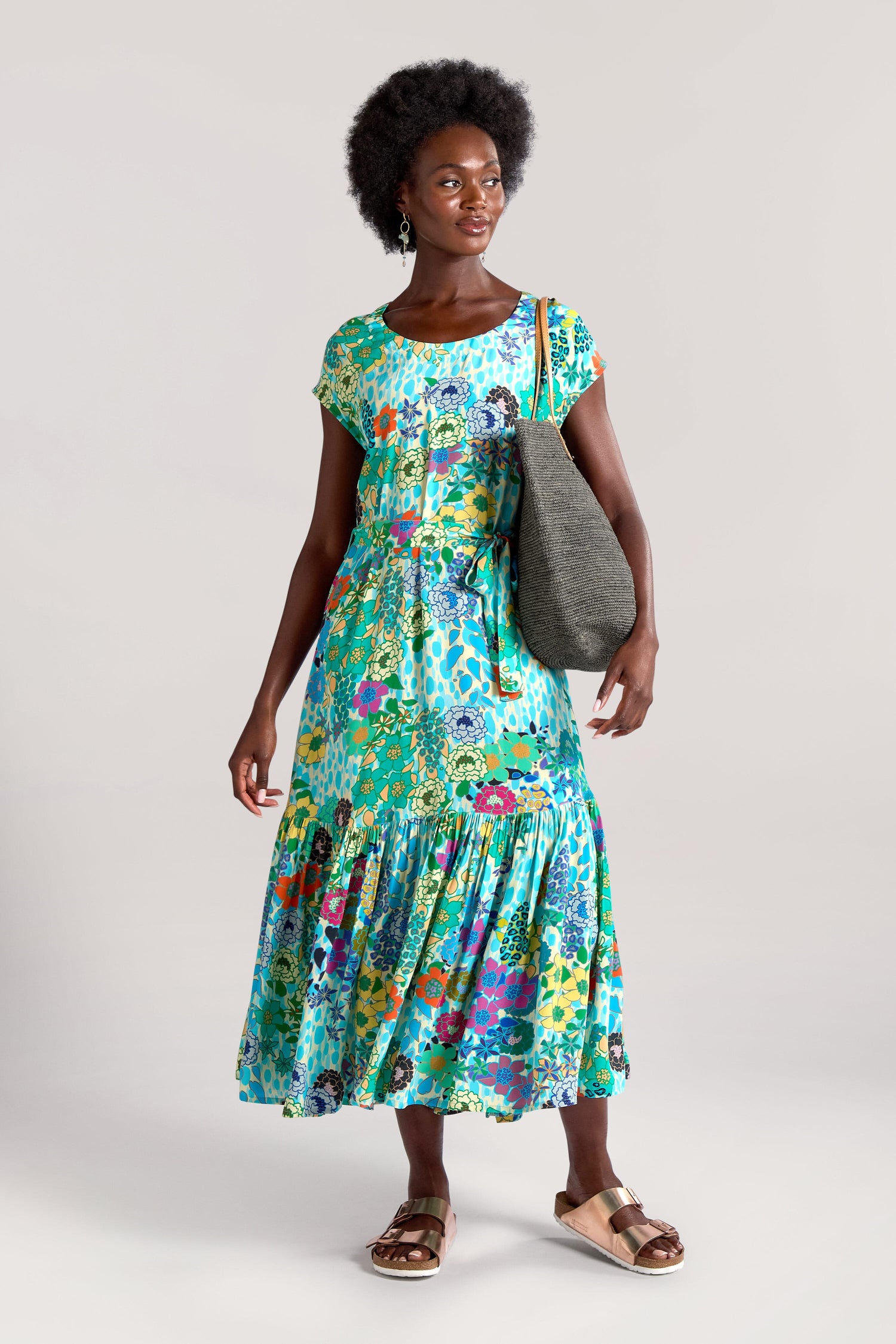 A person wearing a Garden Spot Frill Hem Maxi Dress, gold sandals, and holding a gray tote bag stands against a plain background.