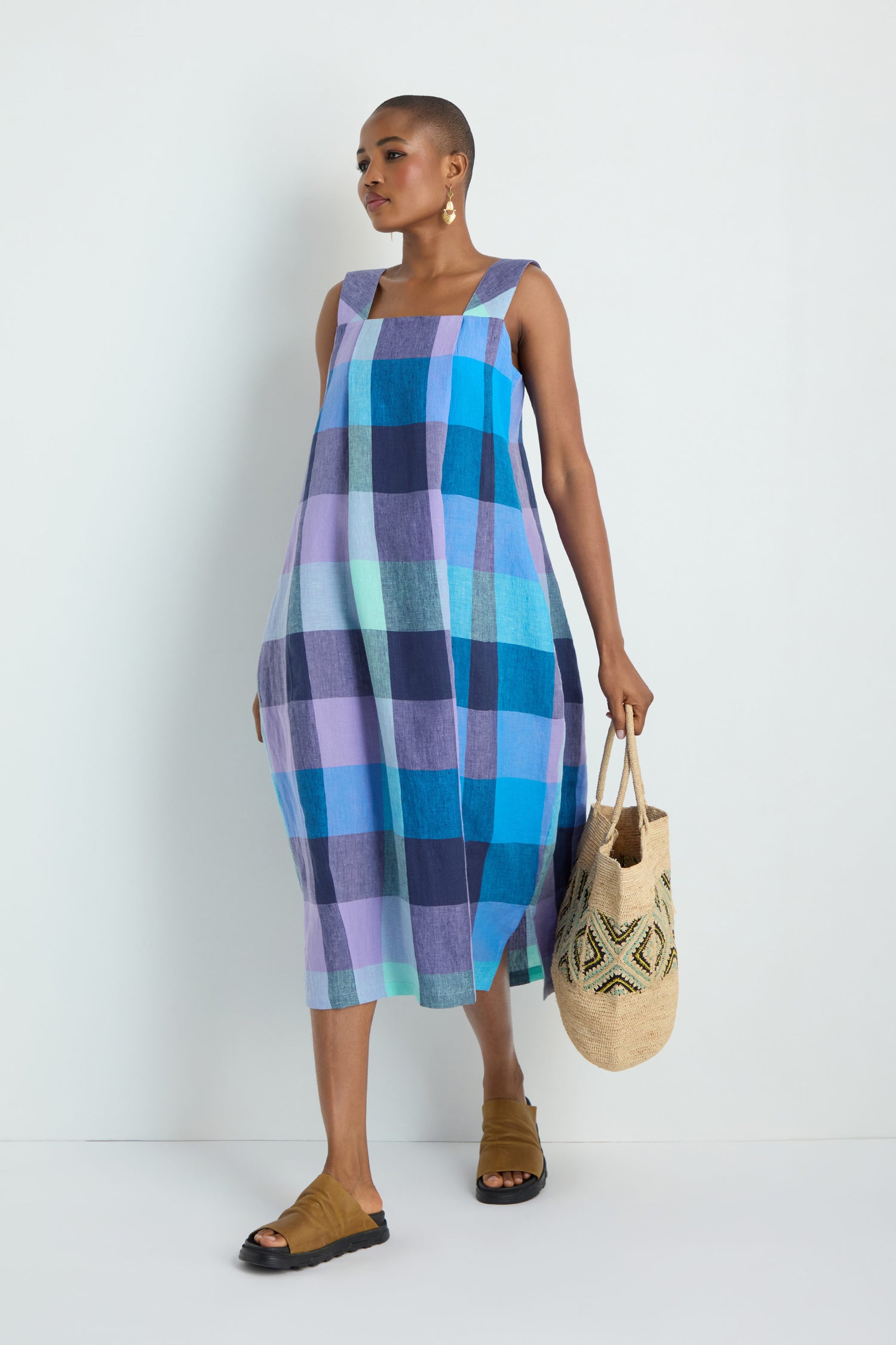 A person wearing the Cross Dye Check Linen Bubble Dress in blue and purple checks, paired with tan sandals and holding a woven tote, stands against a plain white background.