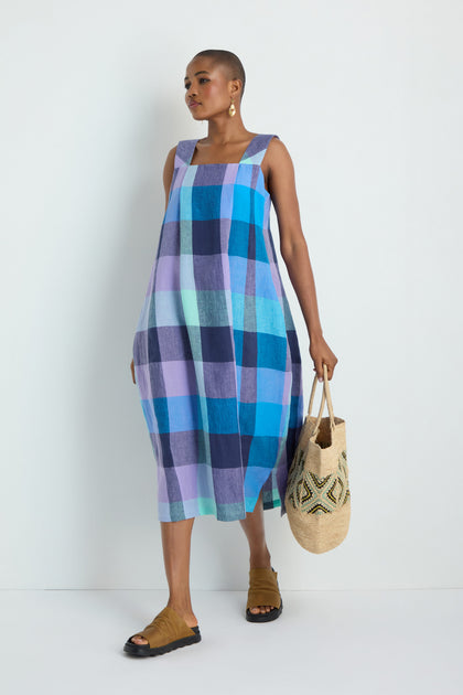 A person wearing the Cross Dye Check Linen Bubble Dress in blue and purple checks, paired with tan sandals and holding a woven tote, stands against a plain white background.
