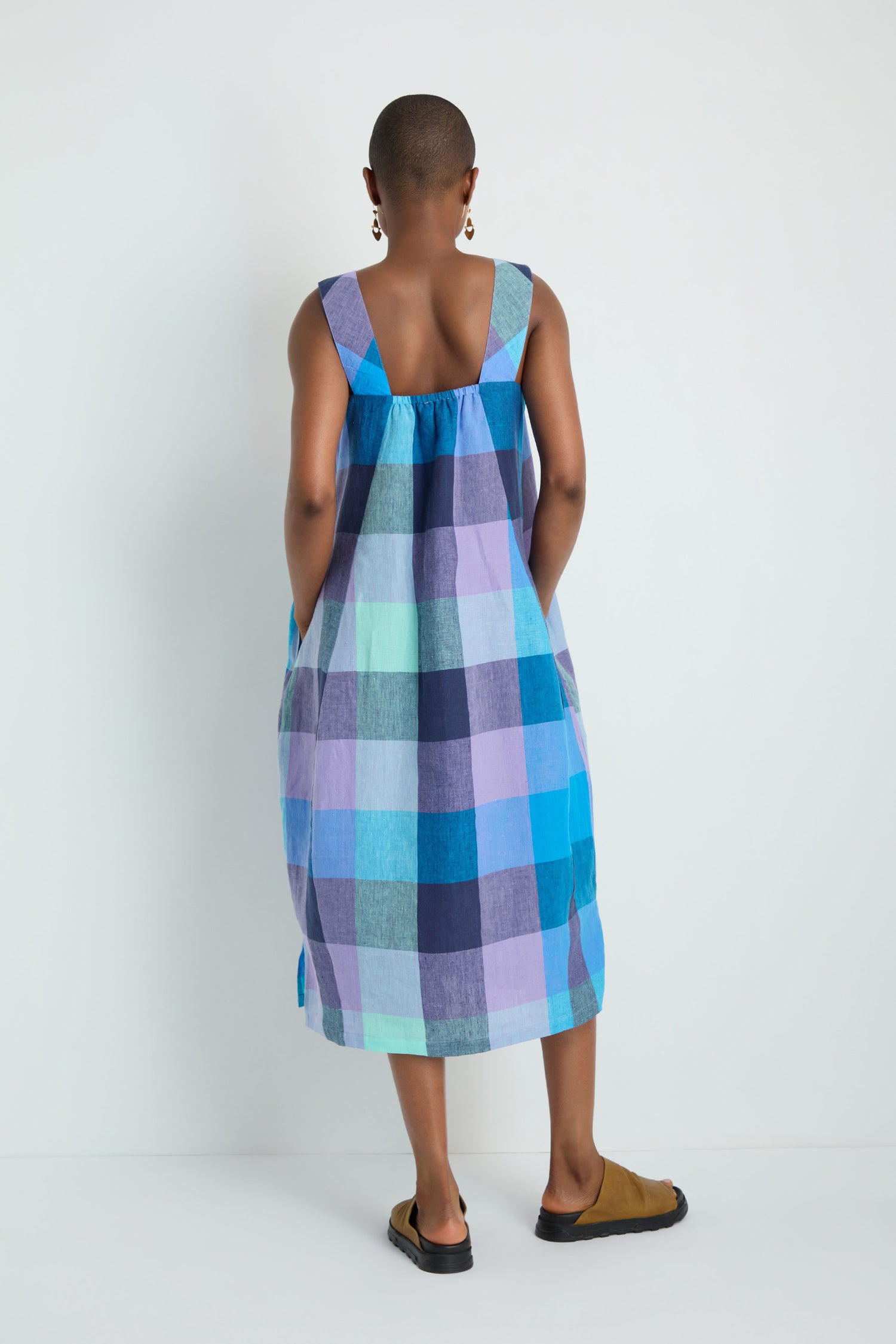 A person with short hair faces away, wearing the Cross Dye Check Linen Bubble Dress in blue and purple plaid yarn-dyed linen, paired with brown sandals and drop earrings against a plain background.