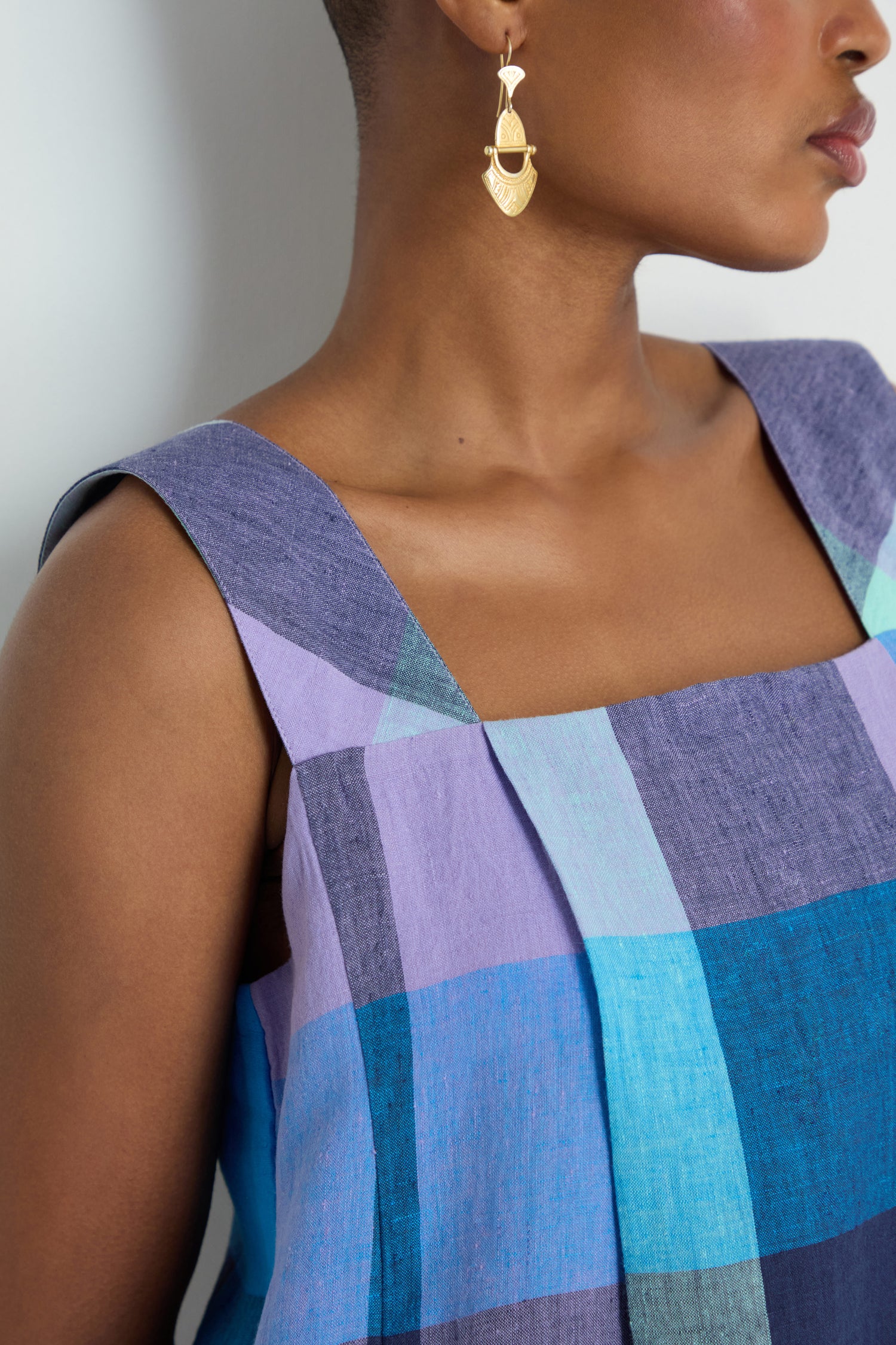 A person wears the Cross Dye Check Linen Bubble Dress in blue and purple yarn-dyed linen with a gold geometric earring, shown from the shoulders up against a plain background.