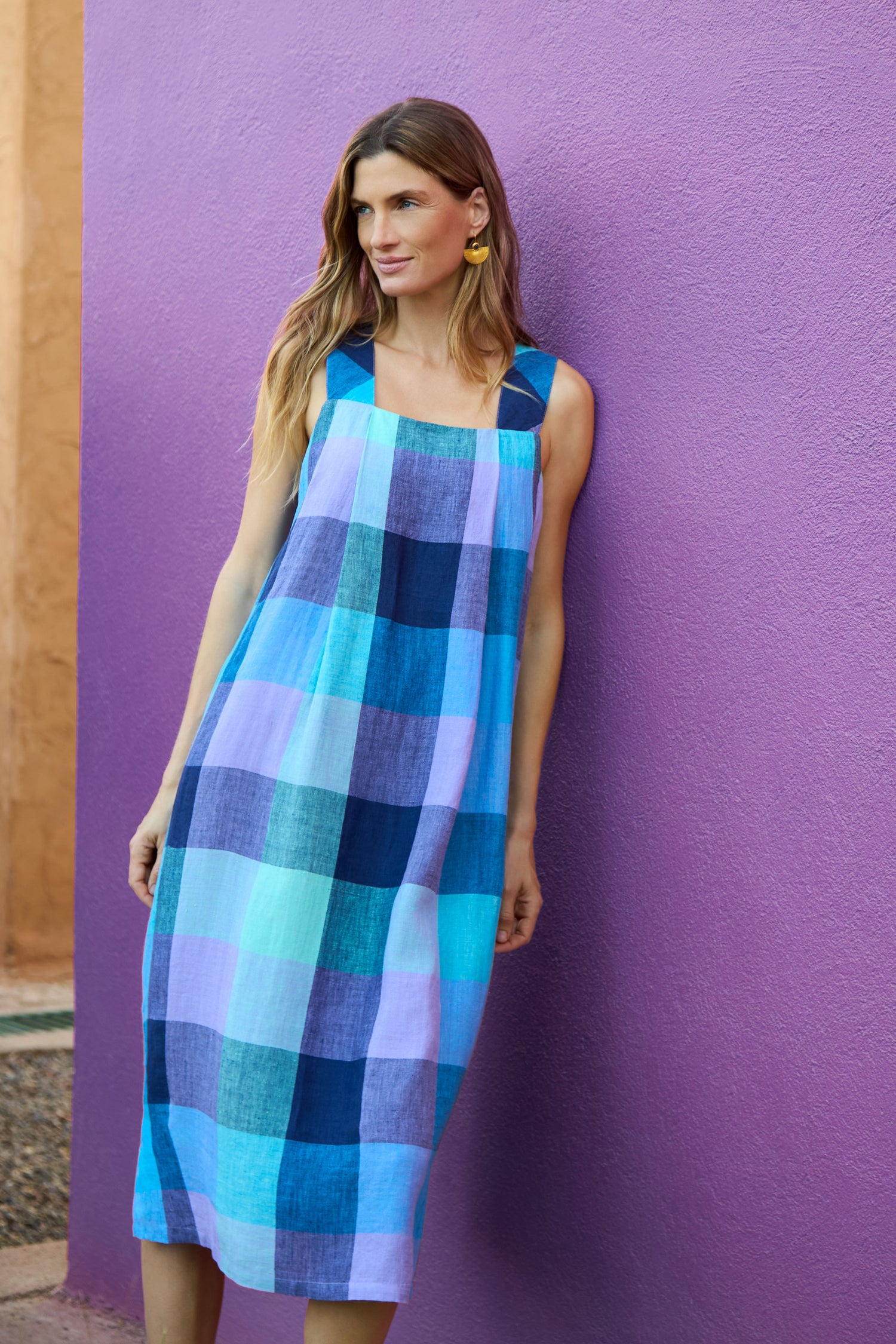 A woman stands against a purple wall wearing the Cross Dye Check Linen Bubble Dress, a sleeveless, loose-fit yarn-dyed linen dress featuring a blue, teal, and purple plaid pattern.